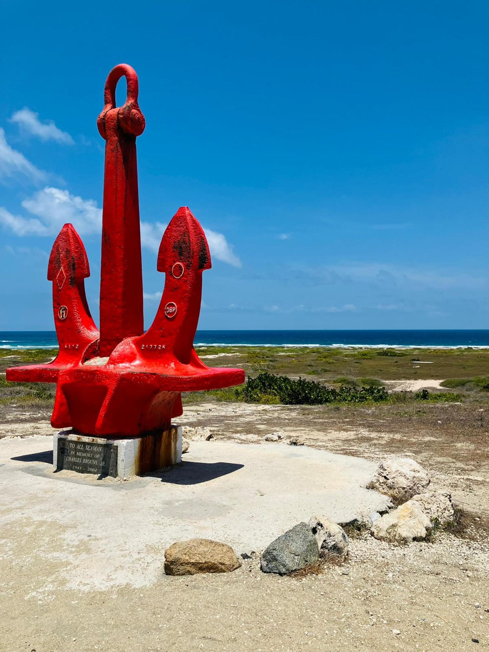 A large red anchor on a concrete base with a plaque, set against a blue sky and coastal landscape.