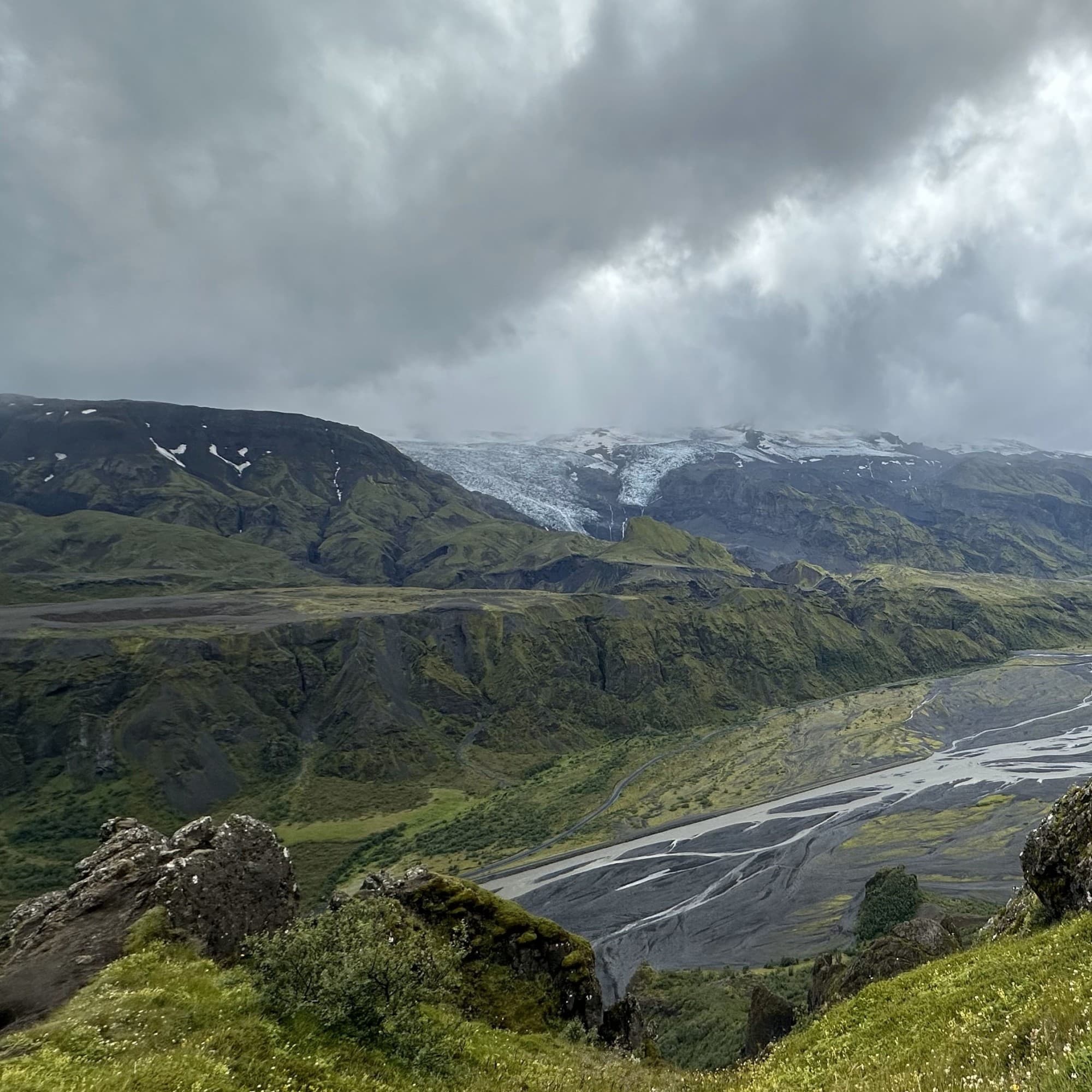 The image shows a panoramic view of a lush, green valley with a meandering river, flanked by mountains and an overcast sky.