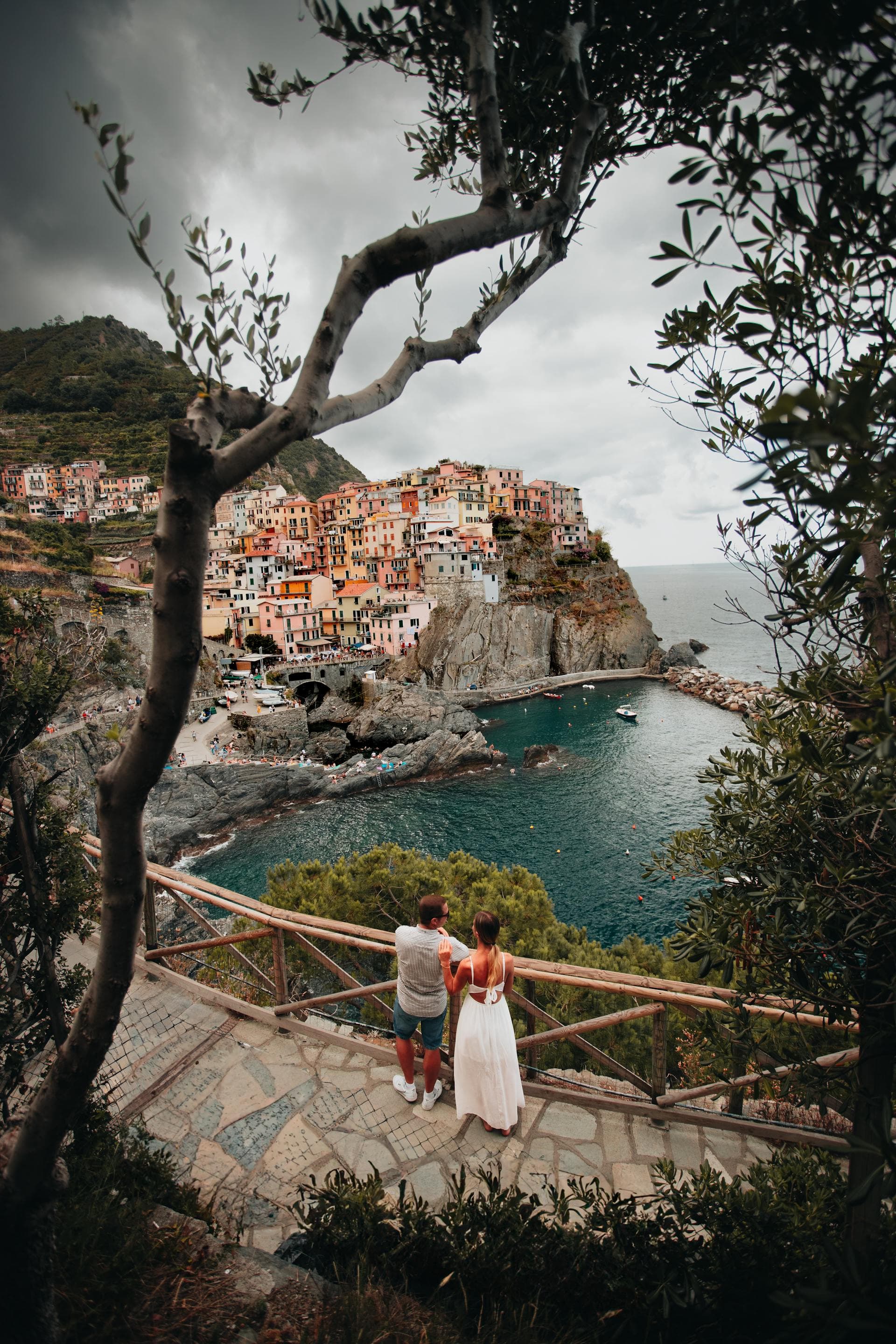 Two people standing by a fence next to the sea with a view of a village atop a hill in the distance