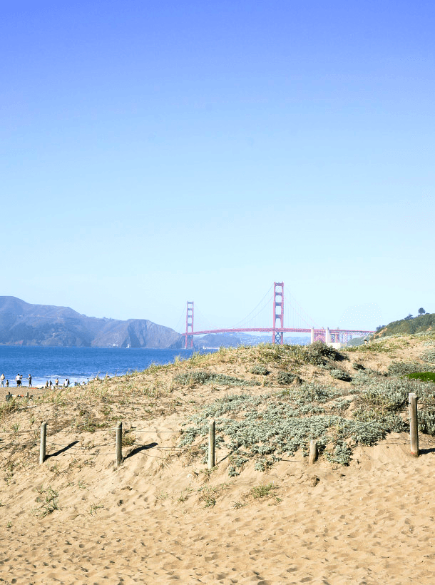 A beach during the daytime with a view of a large, red bridge in the distance