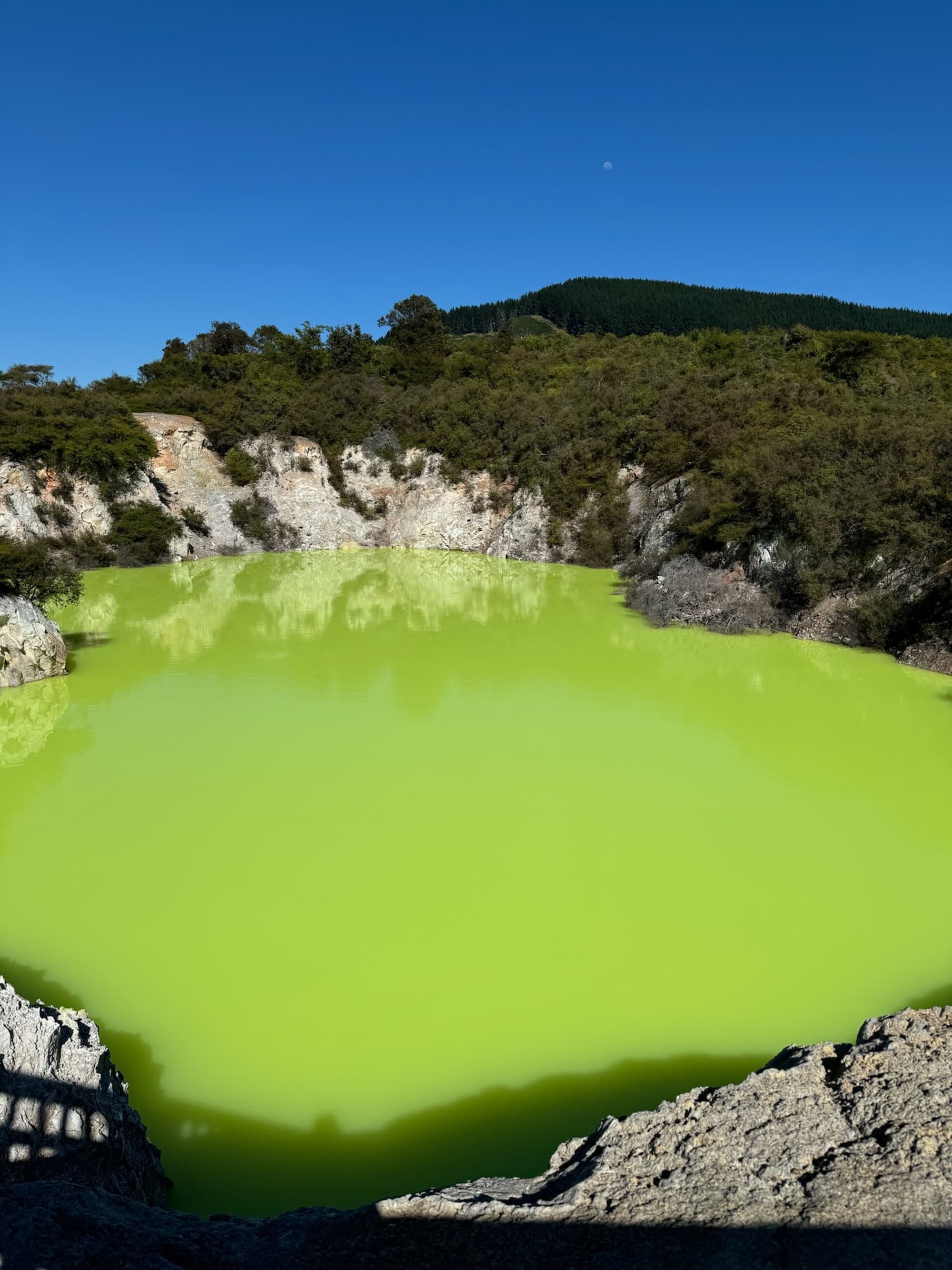 A vibrant green geothermal pool is surrounded by rocky terrain under a clear blue sky.