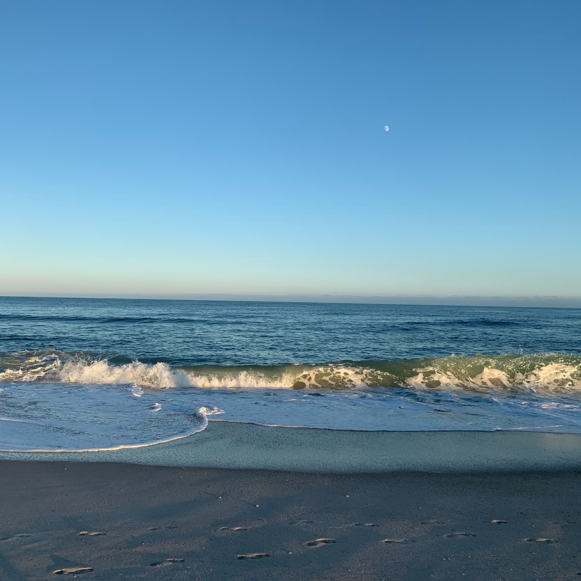 A wave breaks on a sandy beach with clear blue water in the foreground.