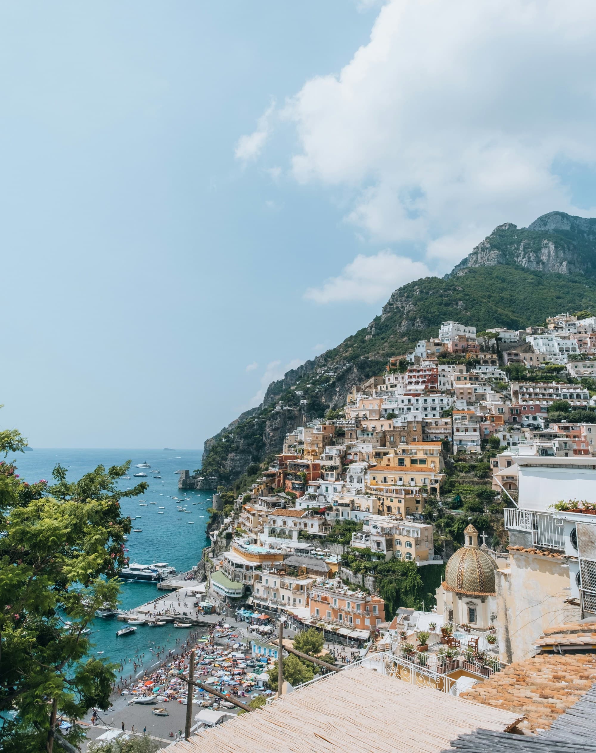 A scenic coastal town with multicolored buildings nestled on a hillside, overlooking a blue sea and beach area under a partly cloudy sky.