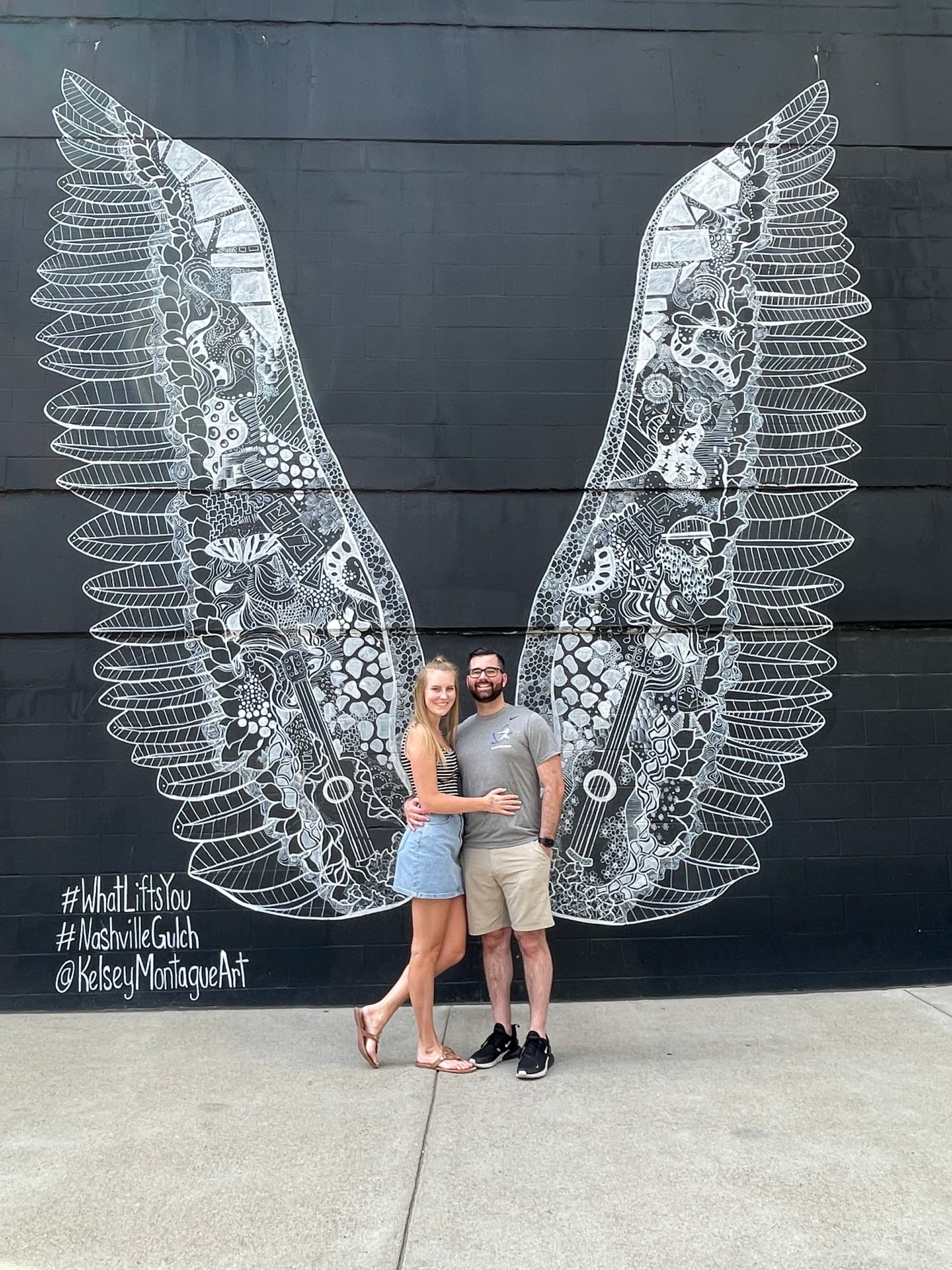 A couple posing for a photo in front of a wall art mural of angel wings