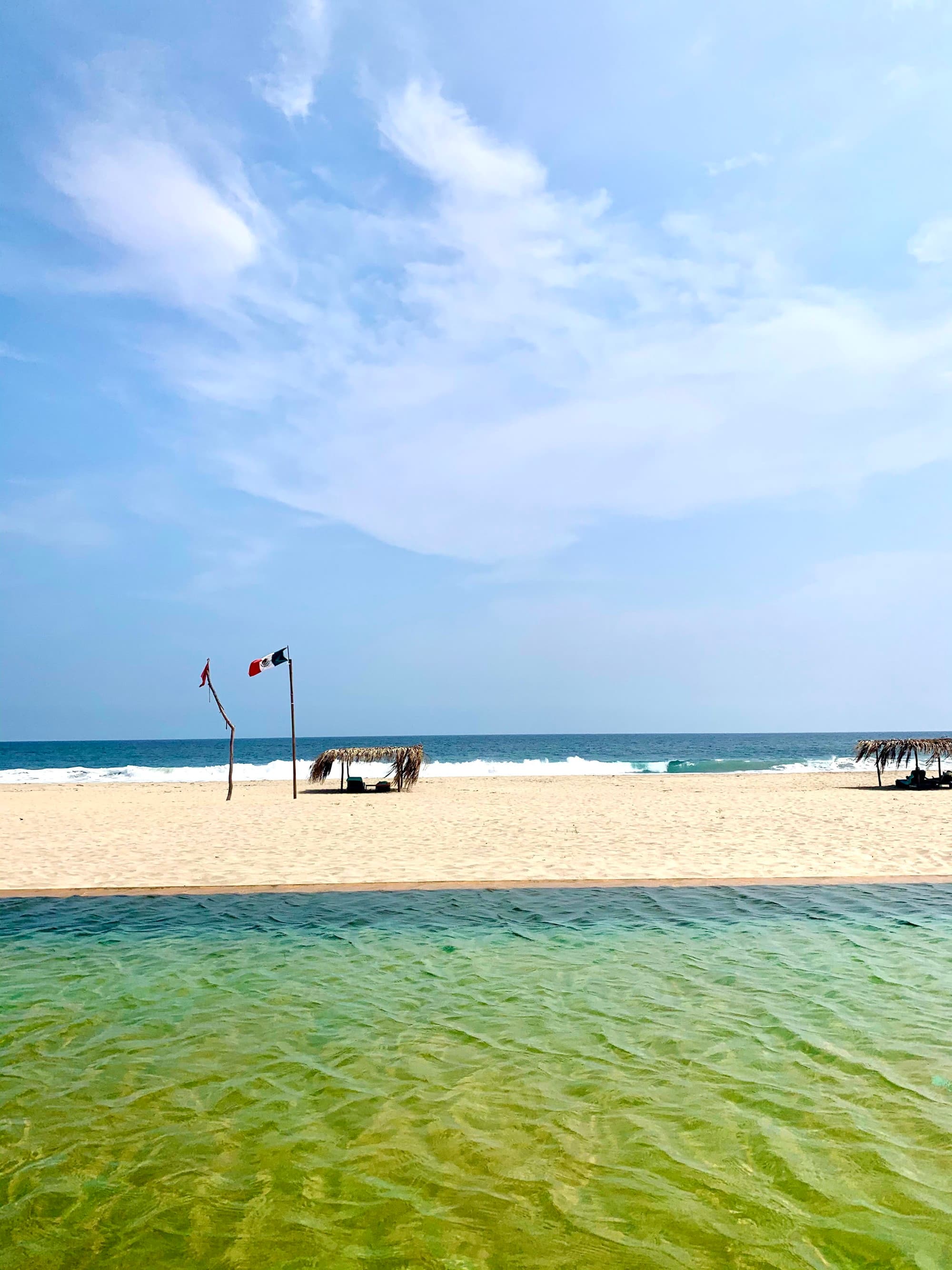 A view of the beach and a small cabana over a chair with the Mexican flag waving in the wind and the ocean washing up on the shore on a partly cloudy day.