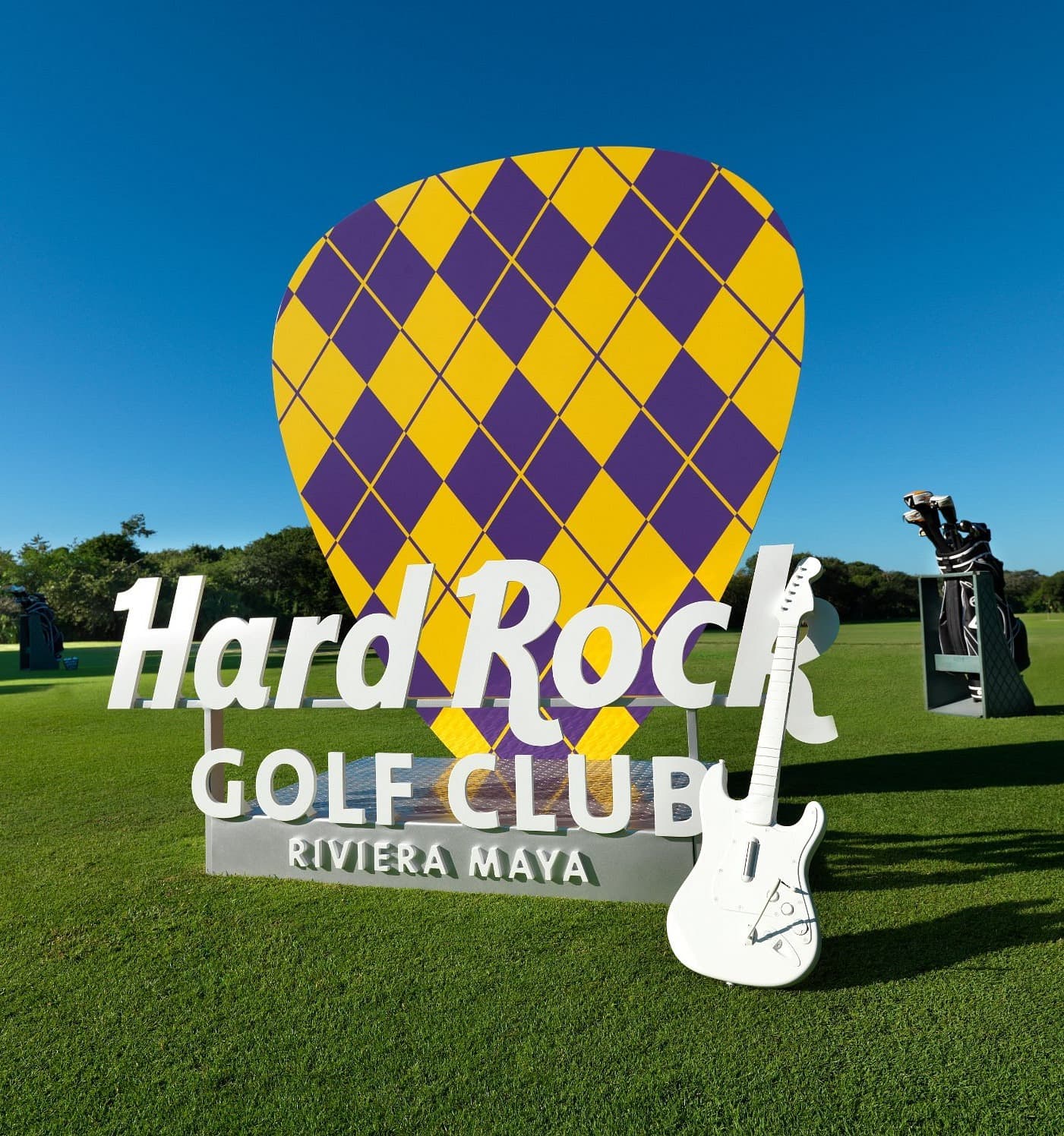 A large sign on a gold course reading "Hard Rock Golf Club Riviera Maya" with a yellow and purple guitar pick statue behind it and a white guitar in front