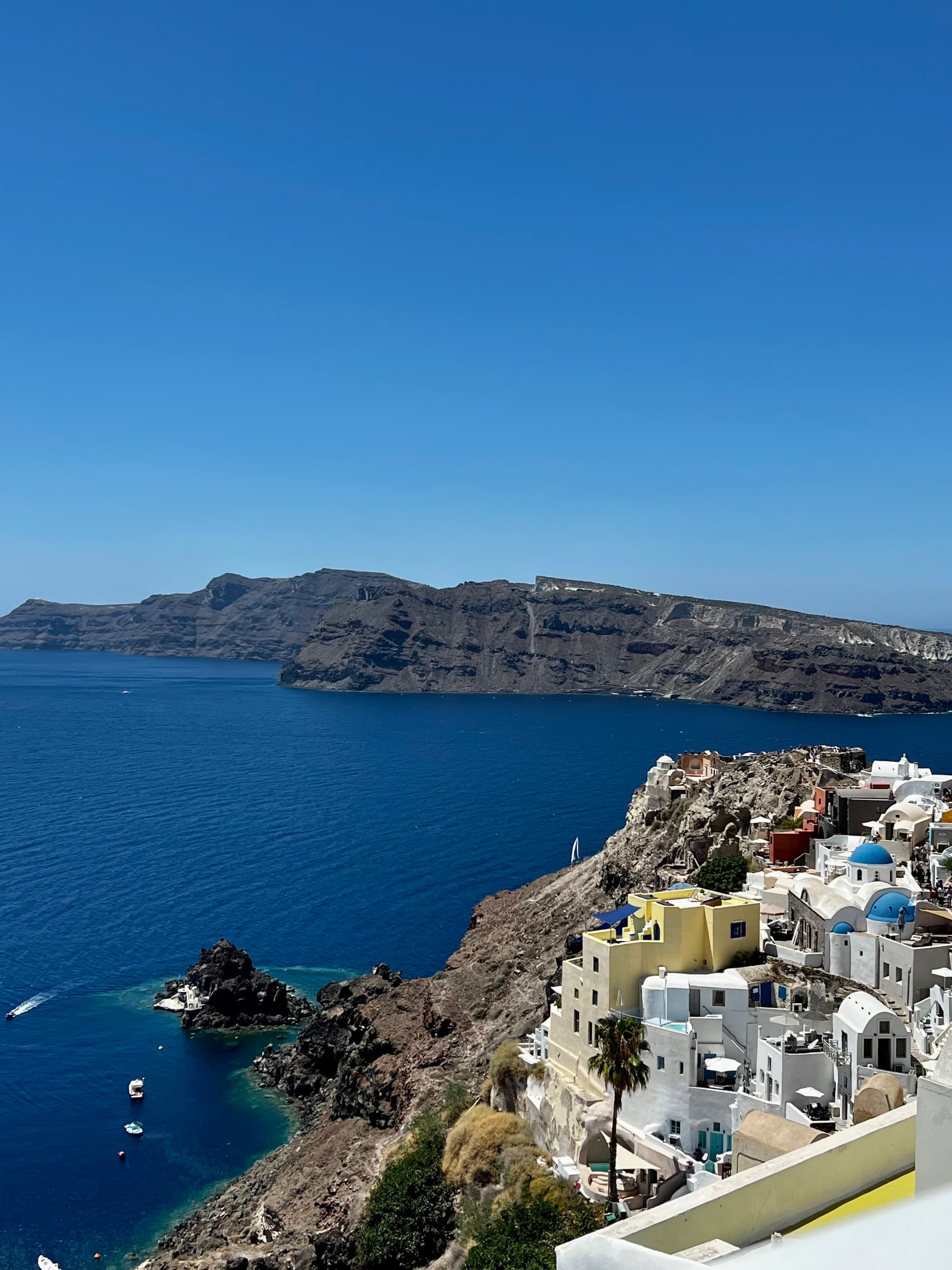 White buildings on a cliffside overlook a blue sea with boats and distant mountains under a clear sky.