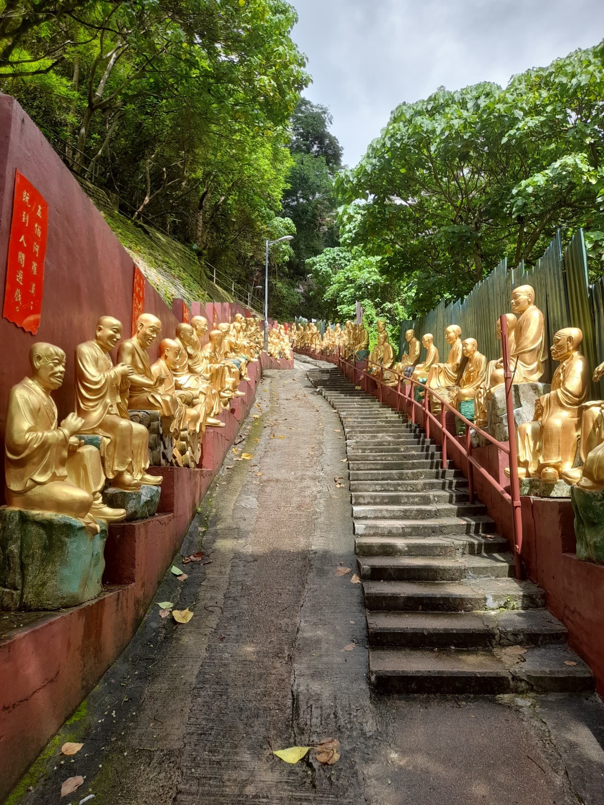 A pathway with stairs lined by golden statues leads up a hill with lush greenery.