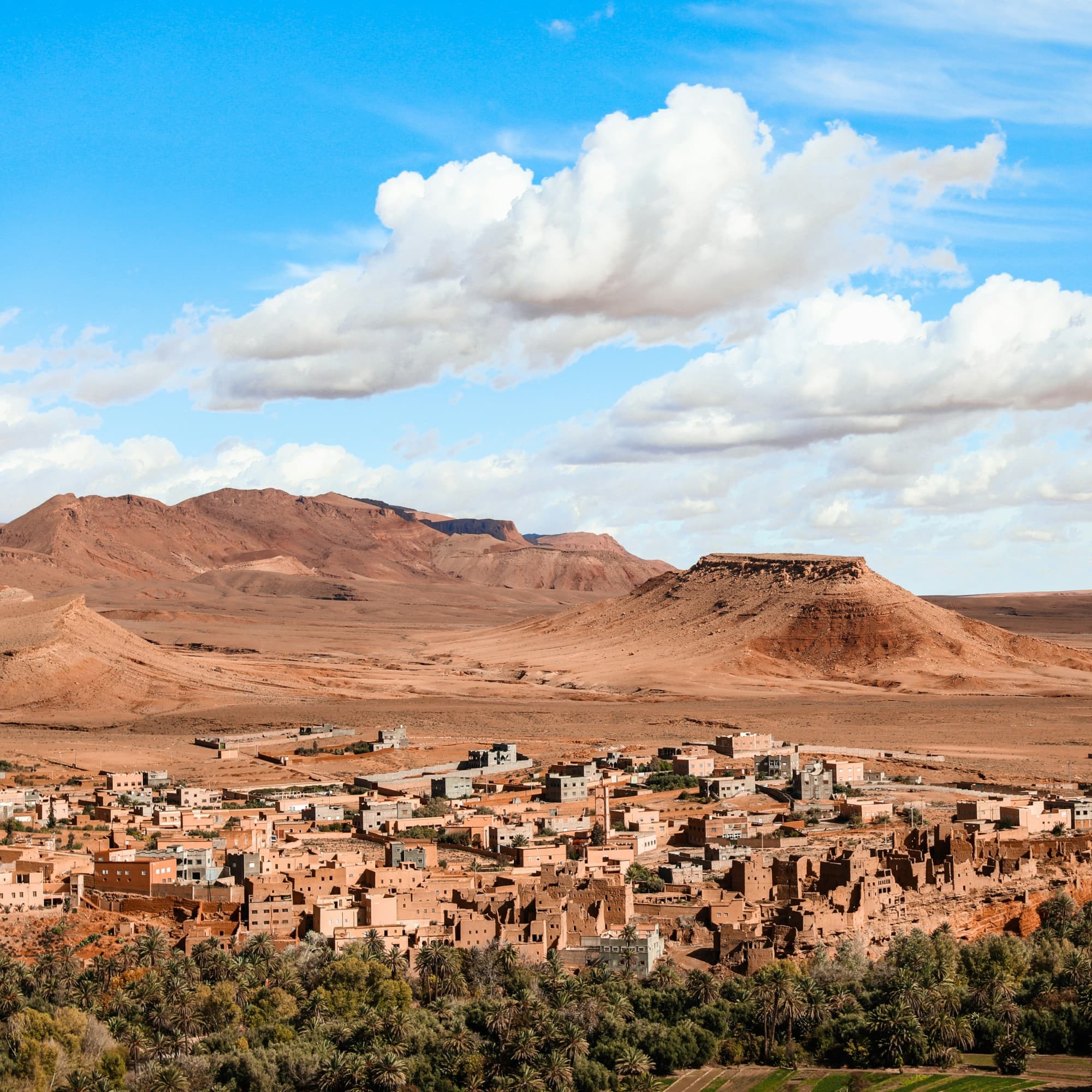 A traditional Moroccan village with clay buildings nestled at the foot of a hill under a blue sky with scattered clouds.