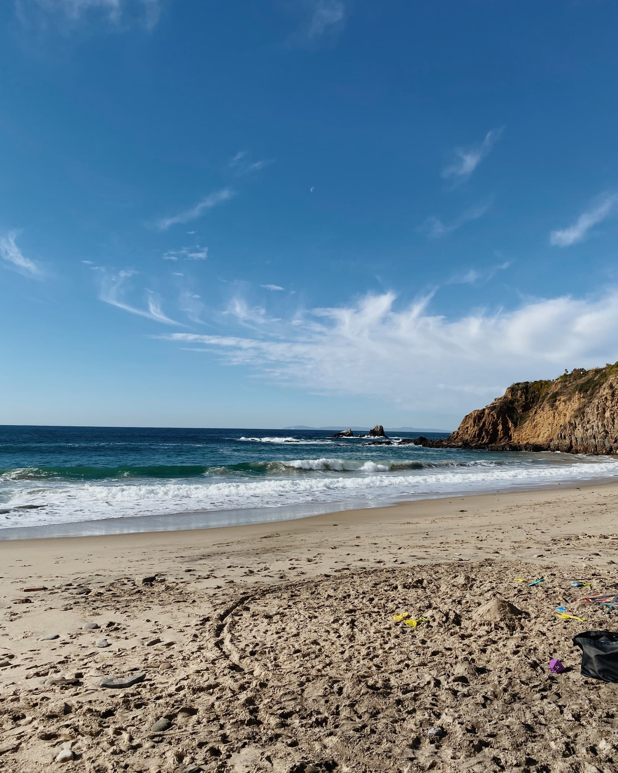 A view of a sandy beach, waves crashing and a cliff in the distance on a clear blue day.