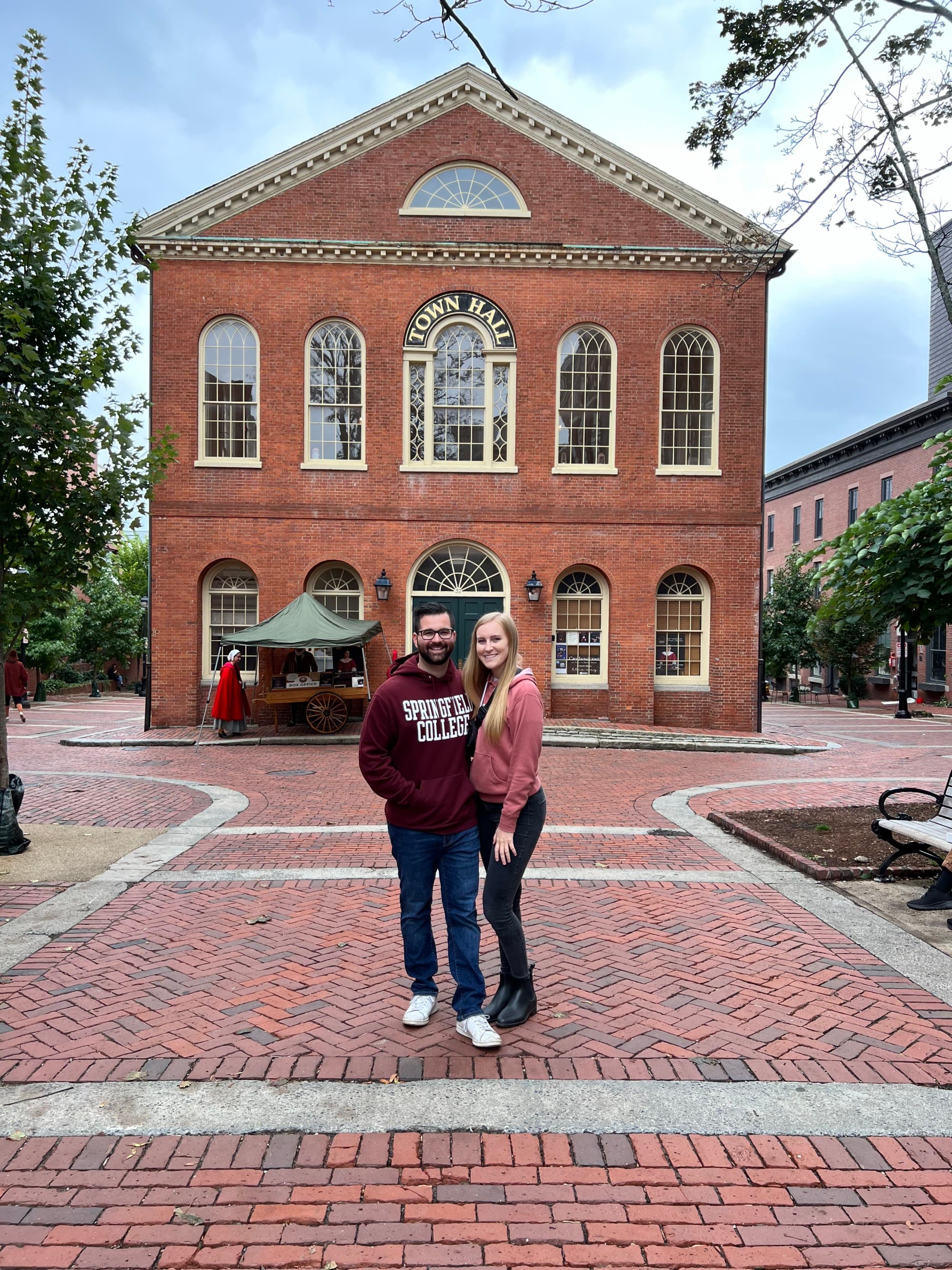 Two individuals pose in front of a brick building with arched windows and a circular window at the top center.