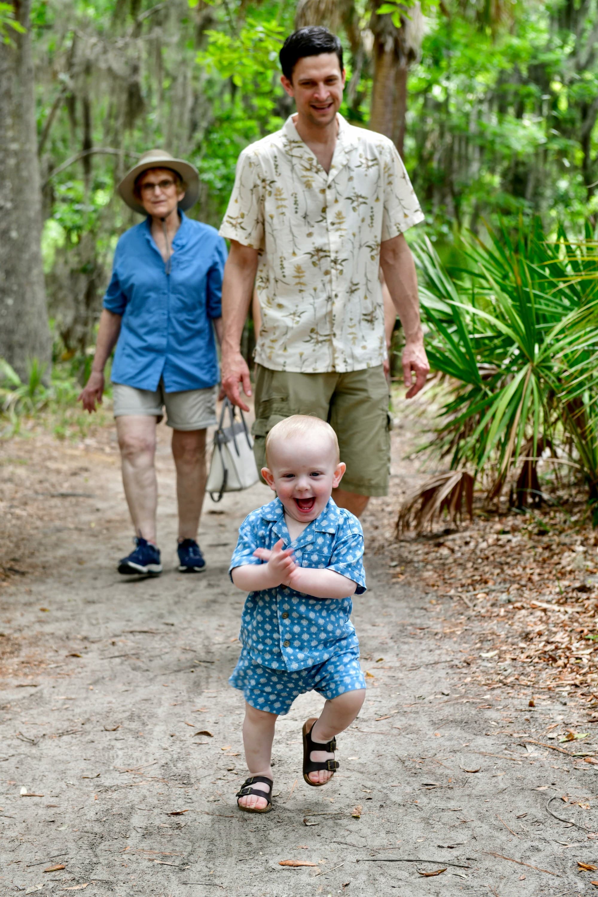 Three people, including a toddler, are walking on a path in a tropical forest.