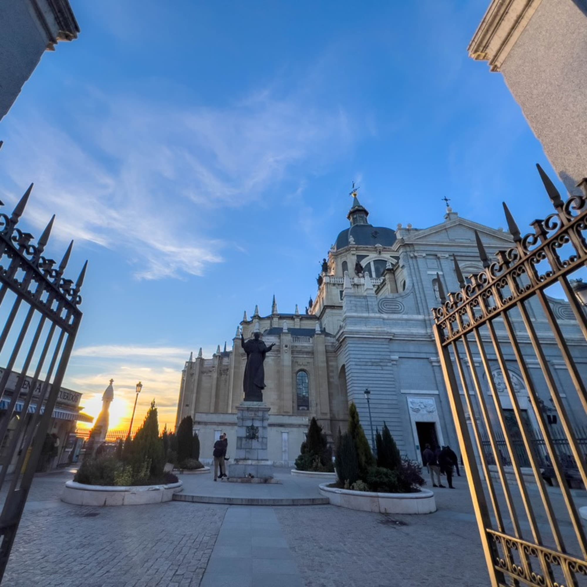 At sunset, An open gate leads to a grand white building with domes under a clear blue sky.