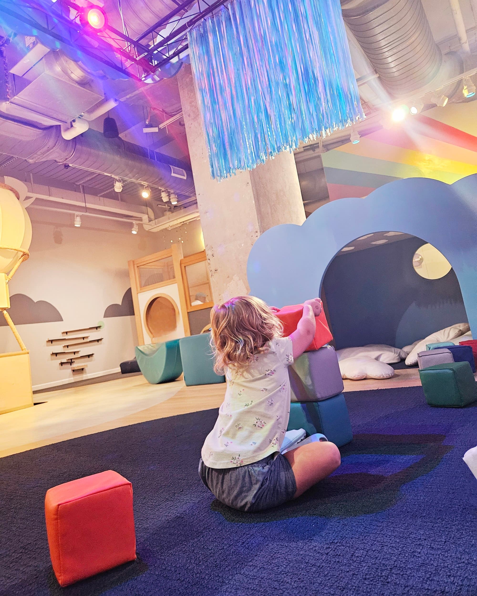 A child enjoying a colorful indoor play area with various structures and soft floor tiles.