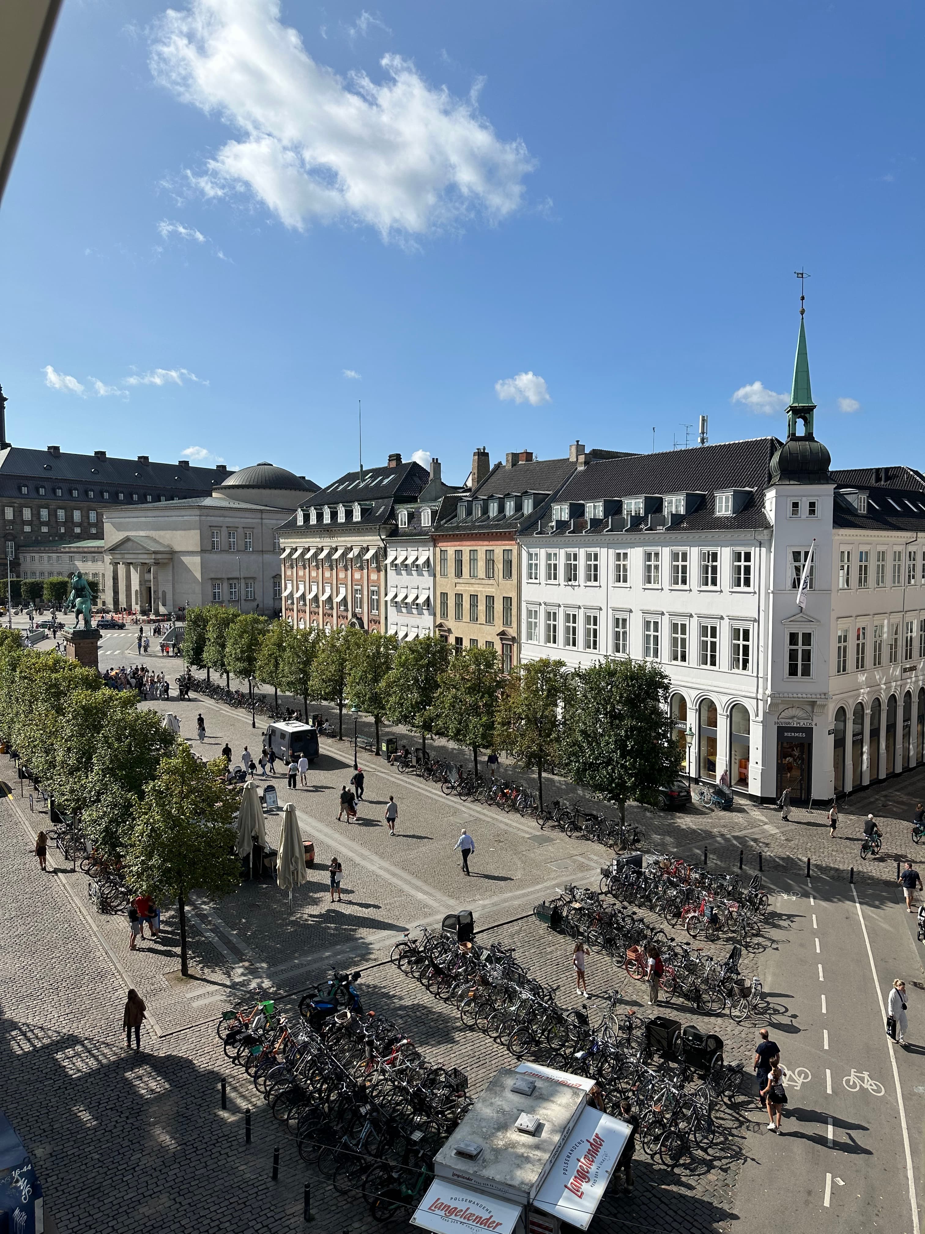 An aerial view of a busy street lined with trees and tall buildings on one side on a sunny day in Copenhagen.