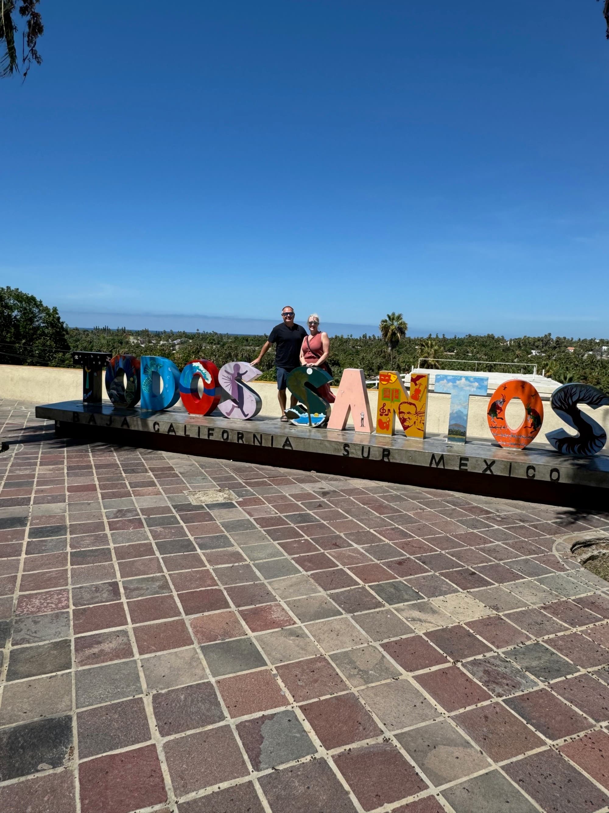 Colorful, decorated Todos Santos Sign under a clear blue sky in an outdoor setting with people standing near it.