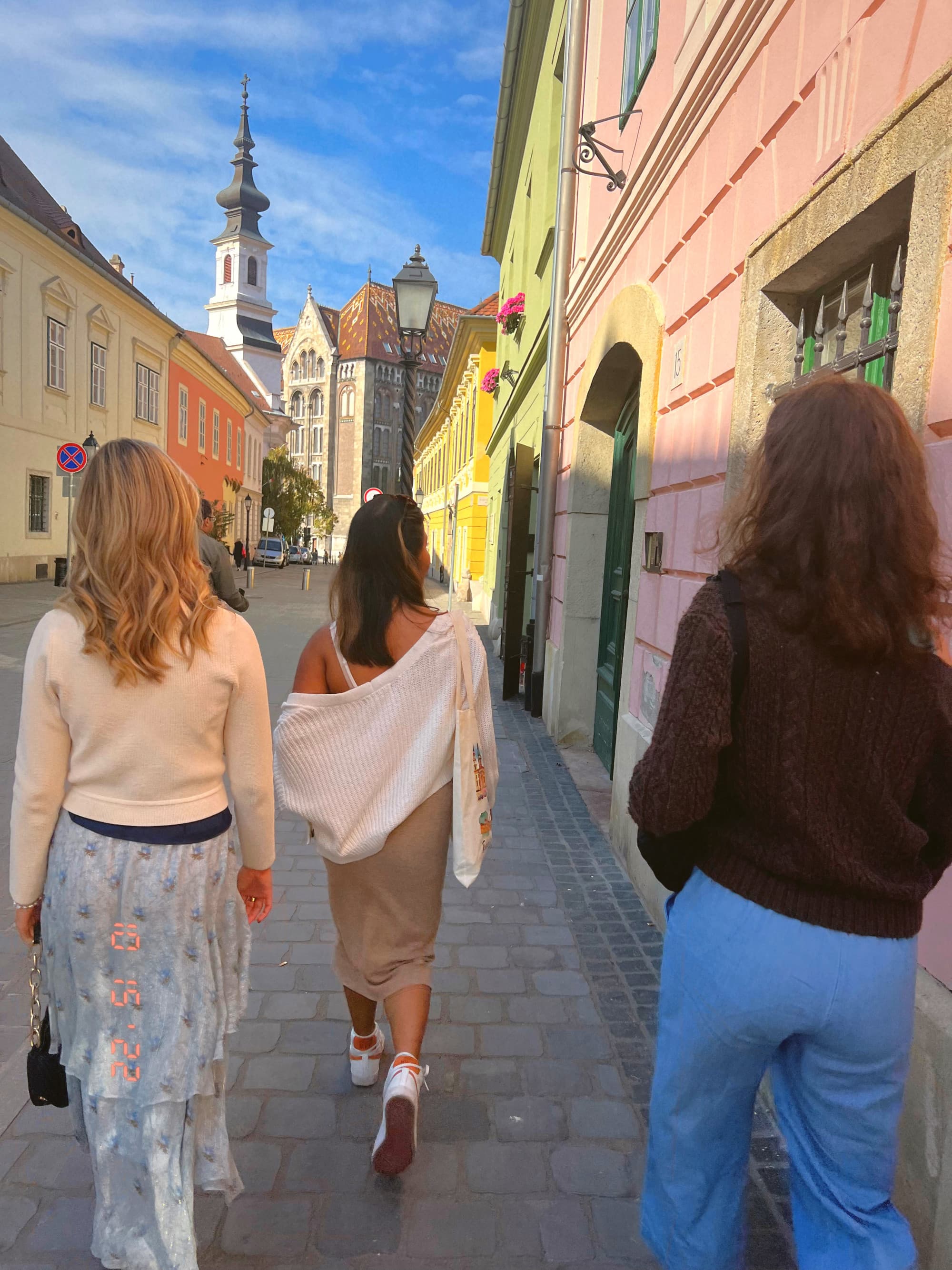 Three people stroll down a charming street with colorful buildings and a church spire in the background.