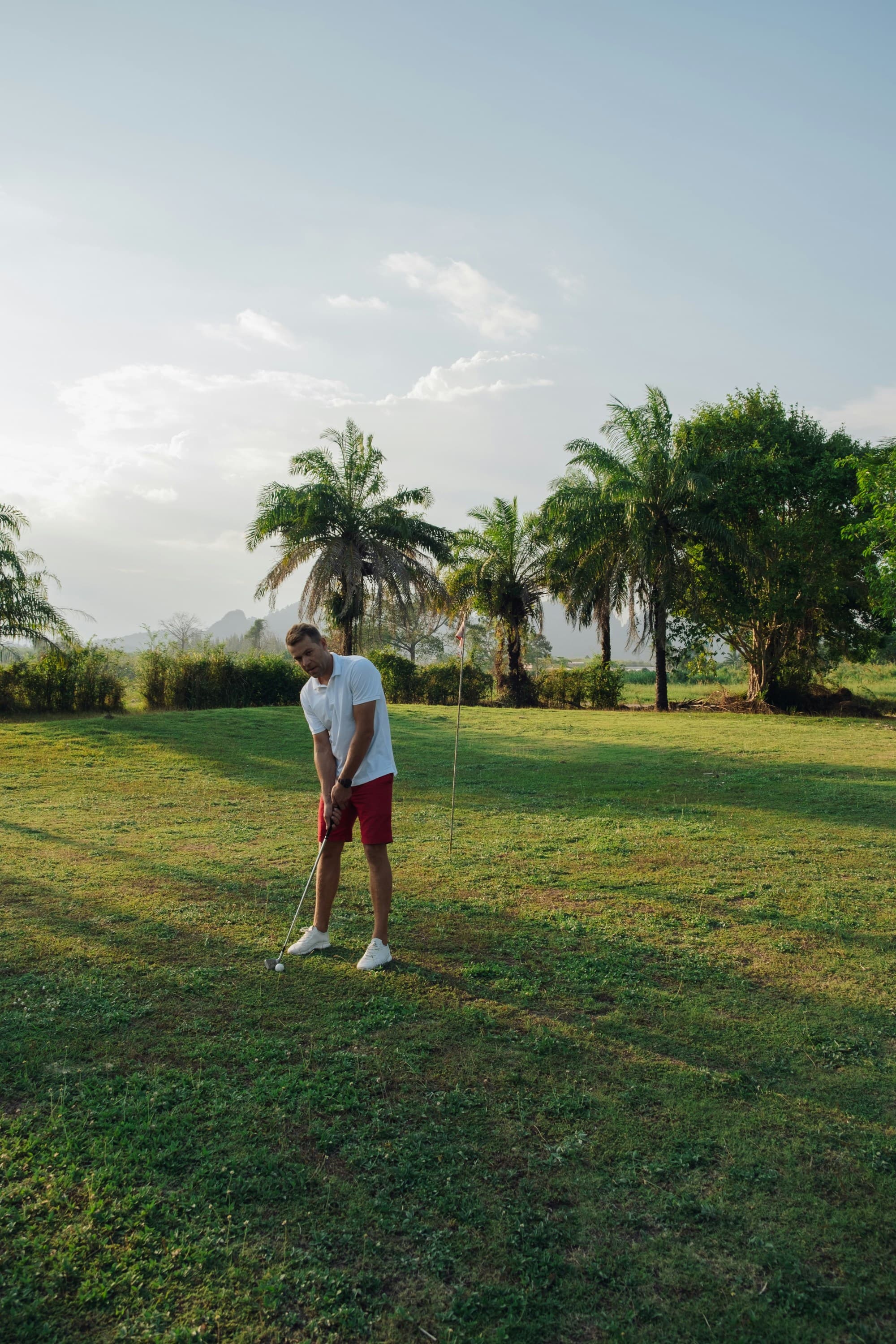 A golfer plays on a lush green course with trees and hills in the background.