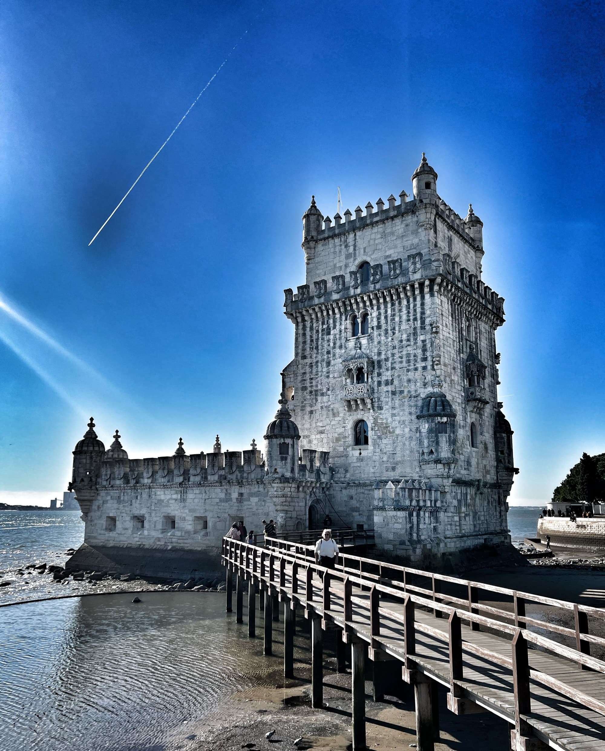 A majestic tower with turrets stands by the water, connected to the shore by a straight pathway.