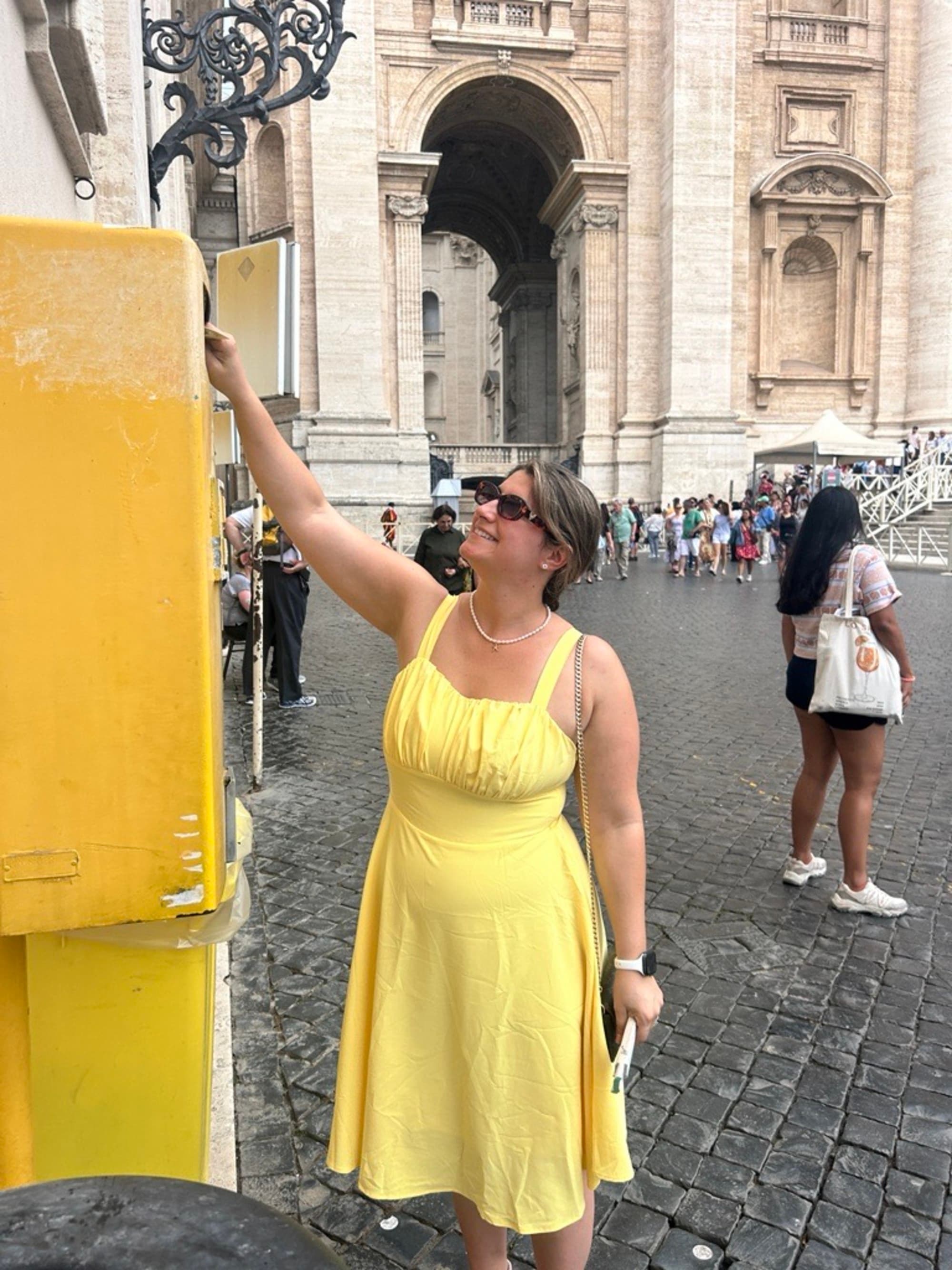 An individual in a yellow dress stands next to a yellow post, with a blurred crowd and historical architecture in the background.