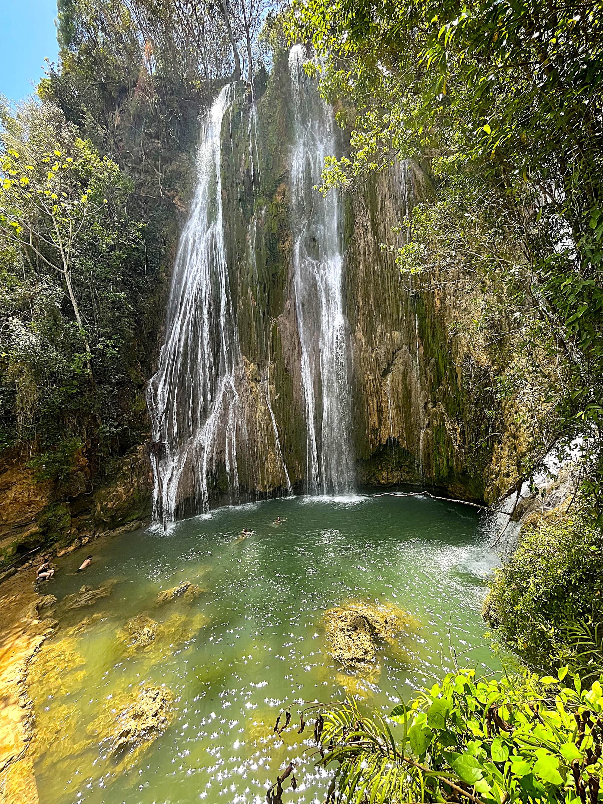 A stunning waterfall cascades into a clear green pool, surrounded by lush greenery on a bright sunny sky.