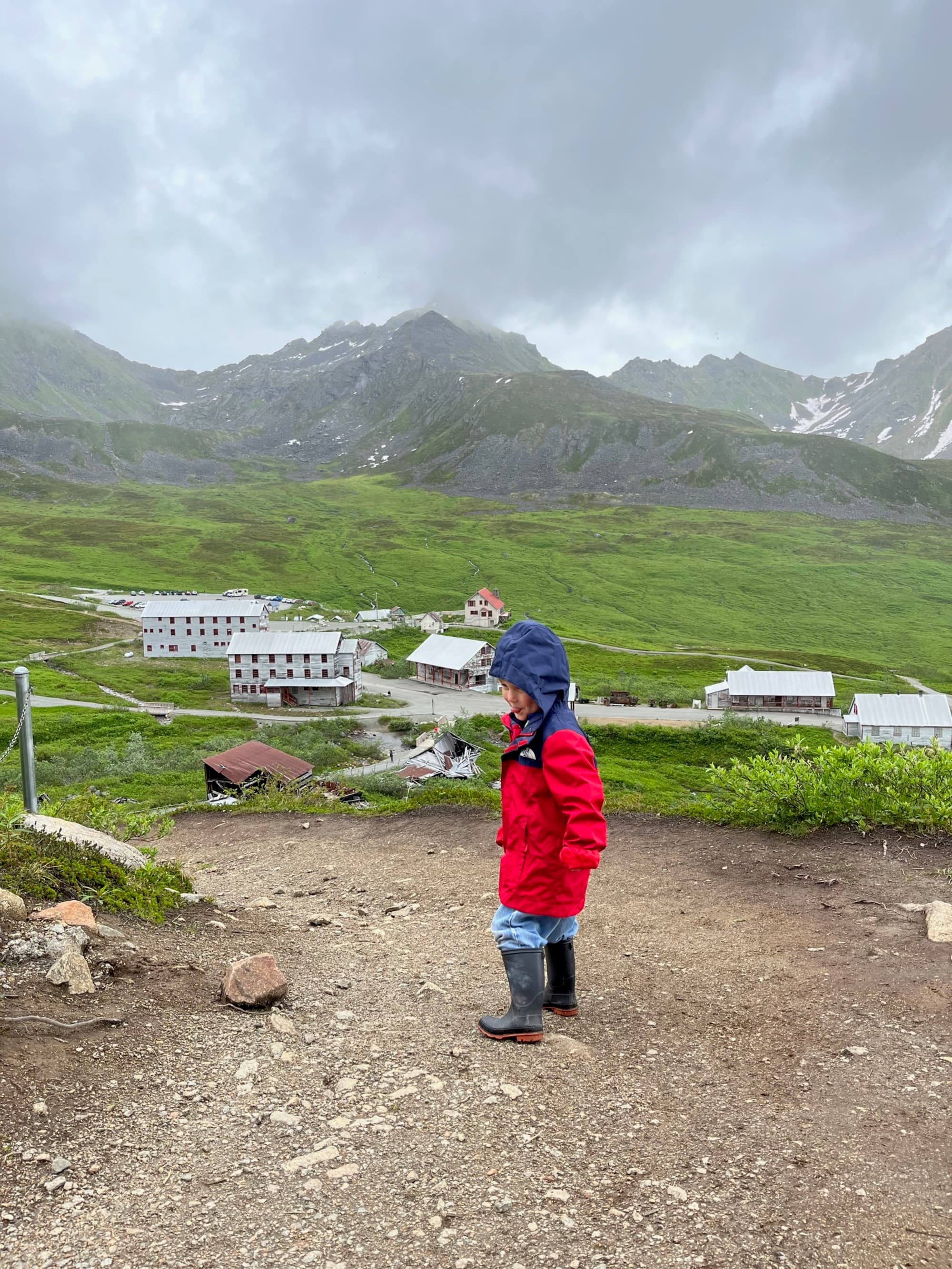 A child in red clothing stands on a mountainous terrain overlooking a valley with buildings, under a cloudy sky.