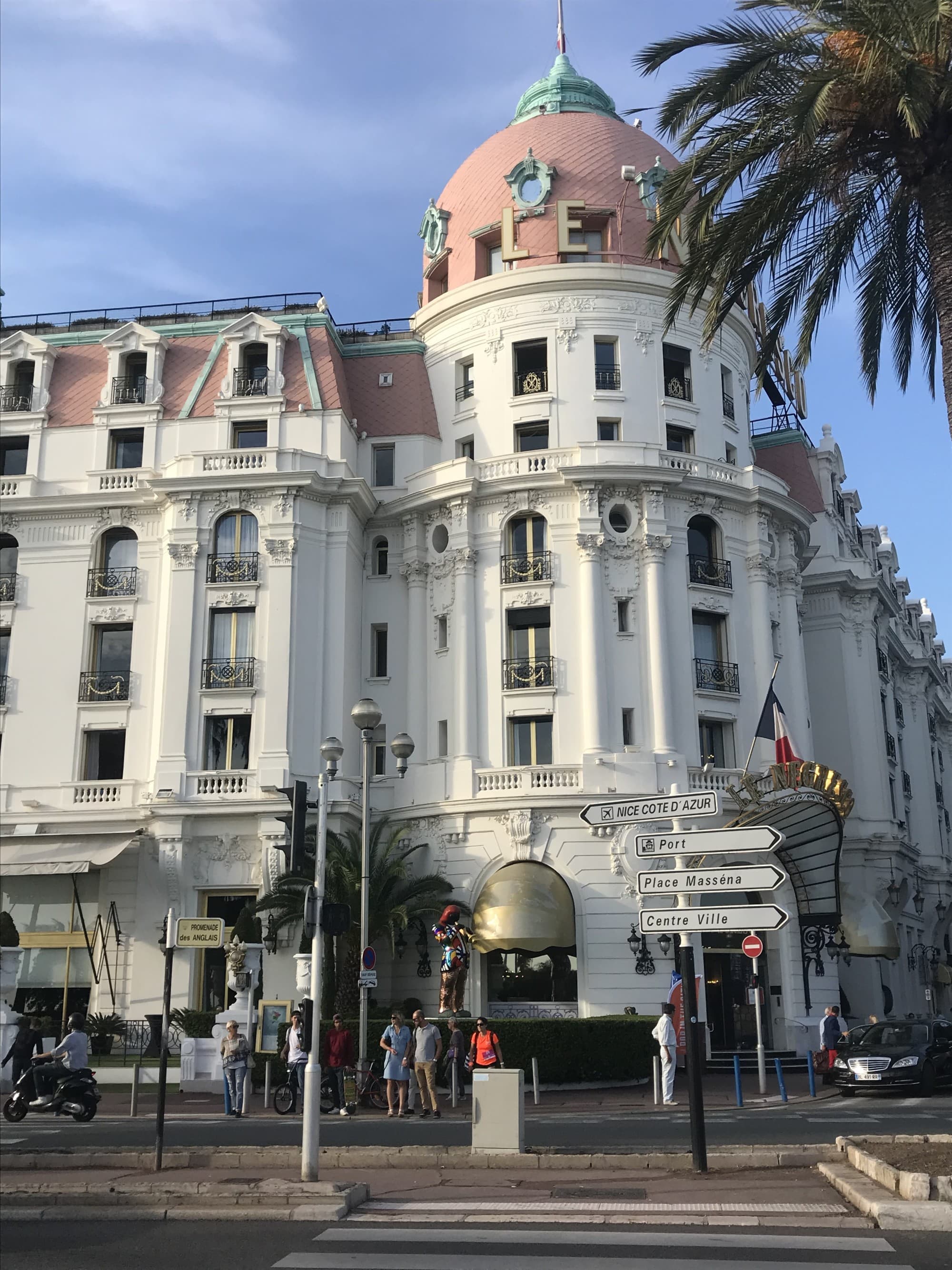 The image shows a grand white building with a green dome and multiple arched windows, situated on a street corner with pedestrians and vehicles nearby.