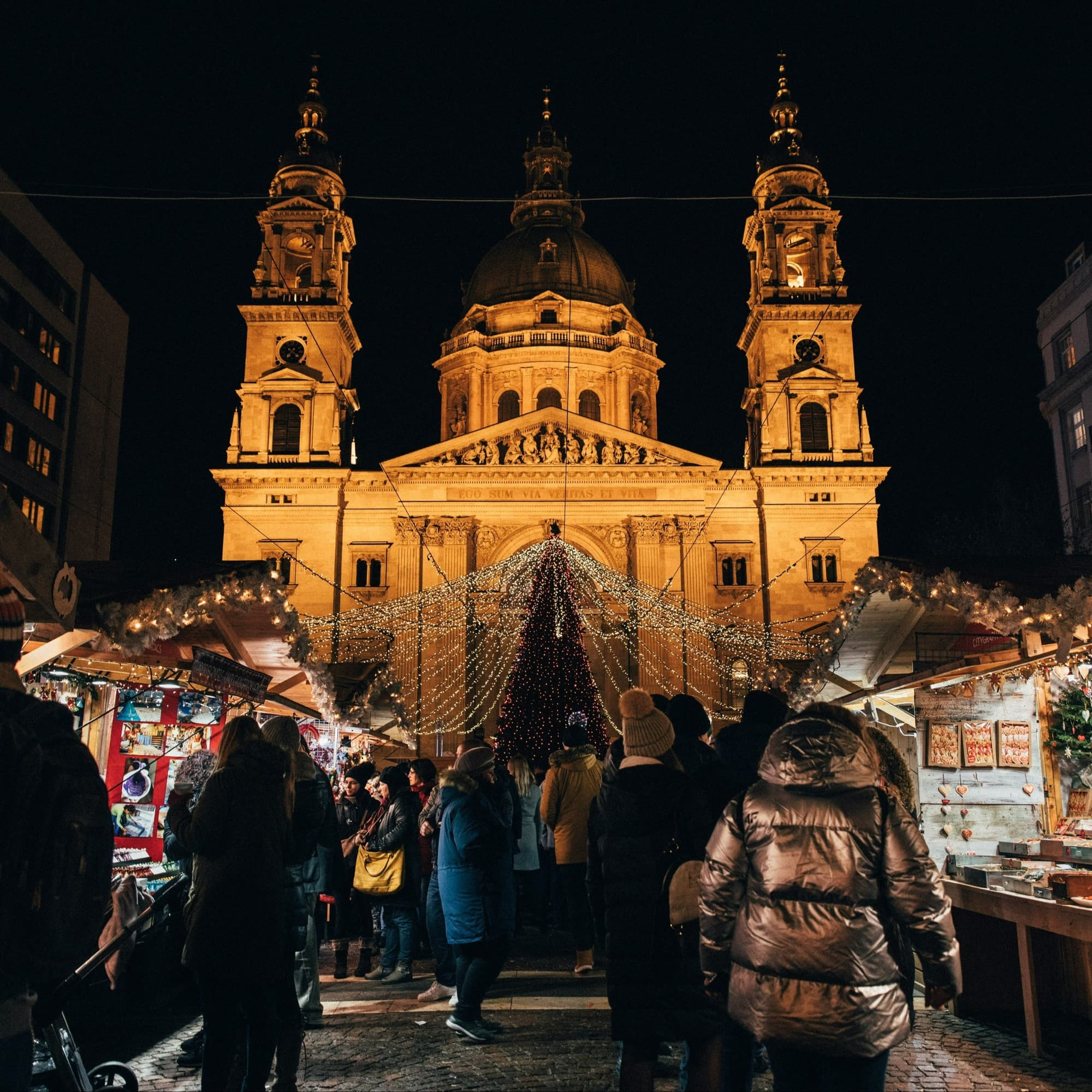 People around well lit stalls at night with a lit up cathedral in the background