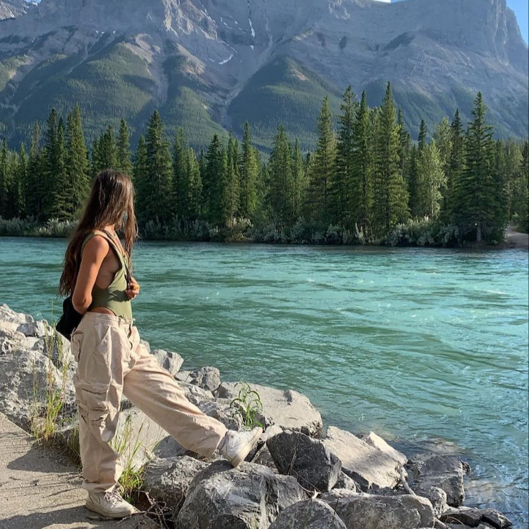 A person stands by a clear blue river, admiring the scenic view of towering mountains and lush green trees.