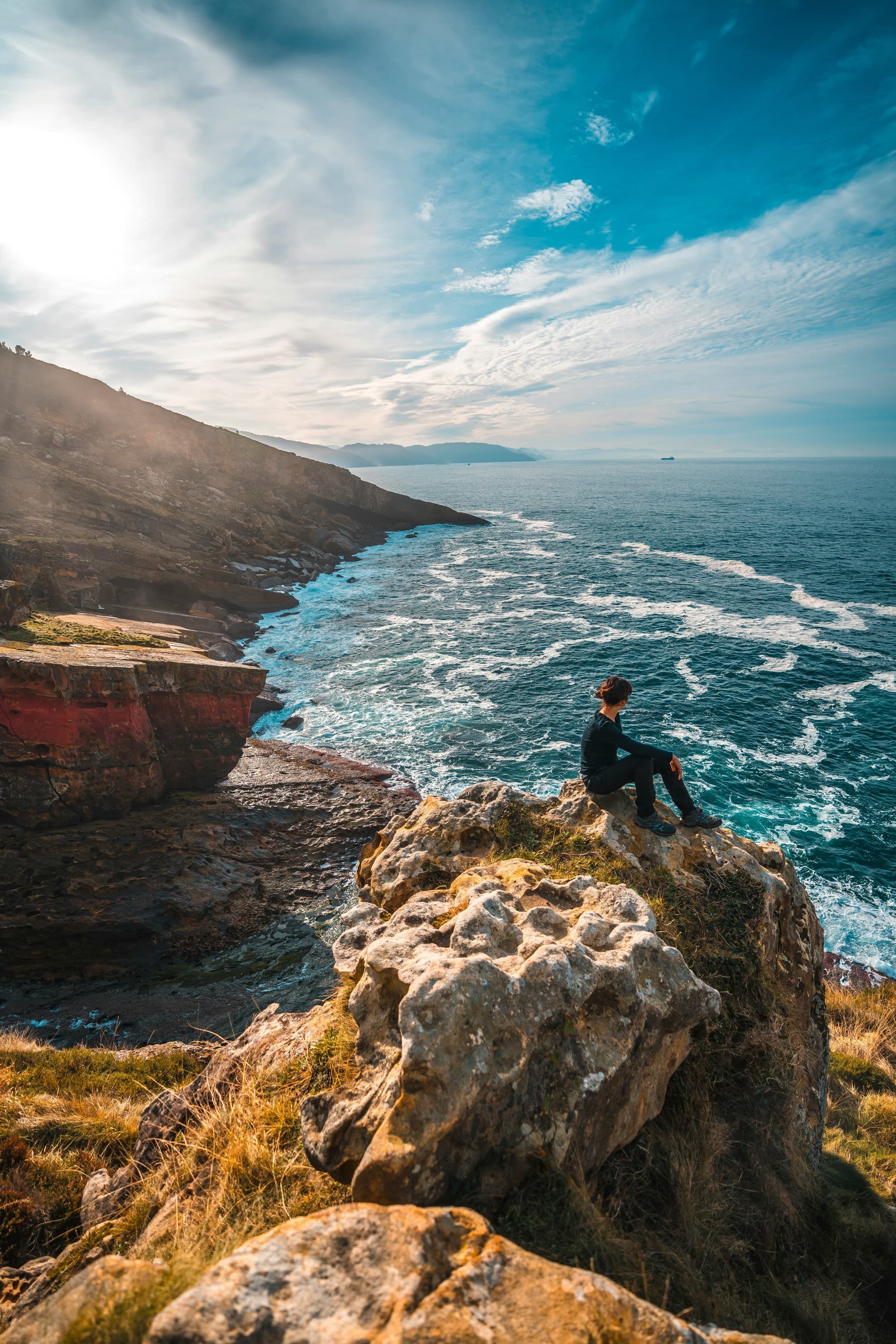 The image depicts an individual sitting on a rocky outcrop overlooking a vibrant blue sea with a clear sky above.