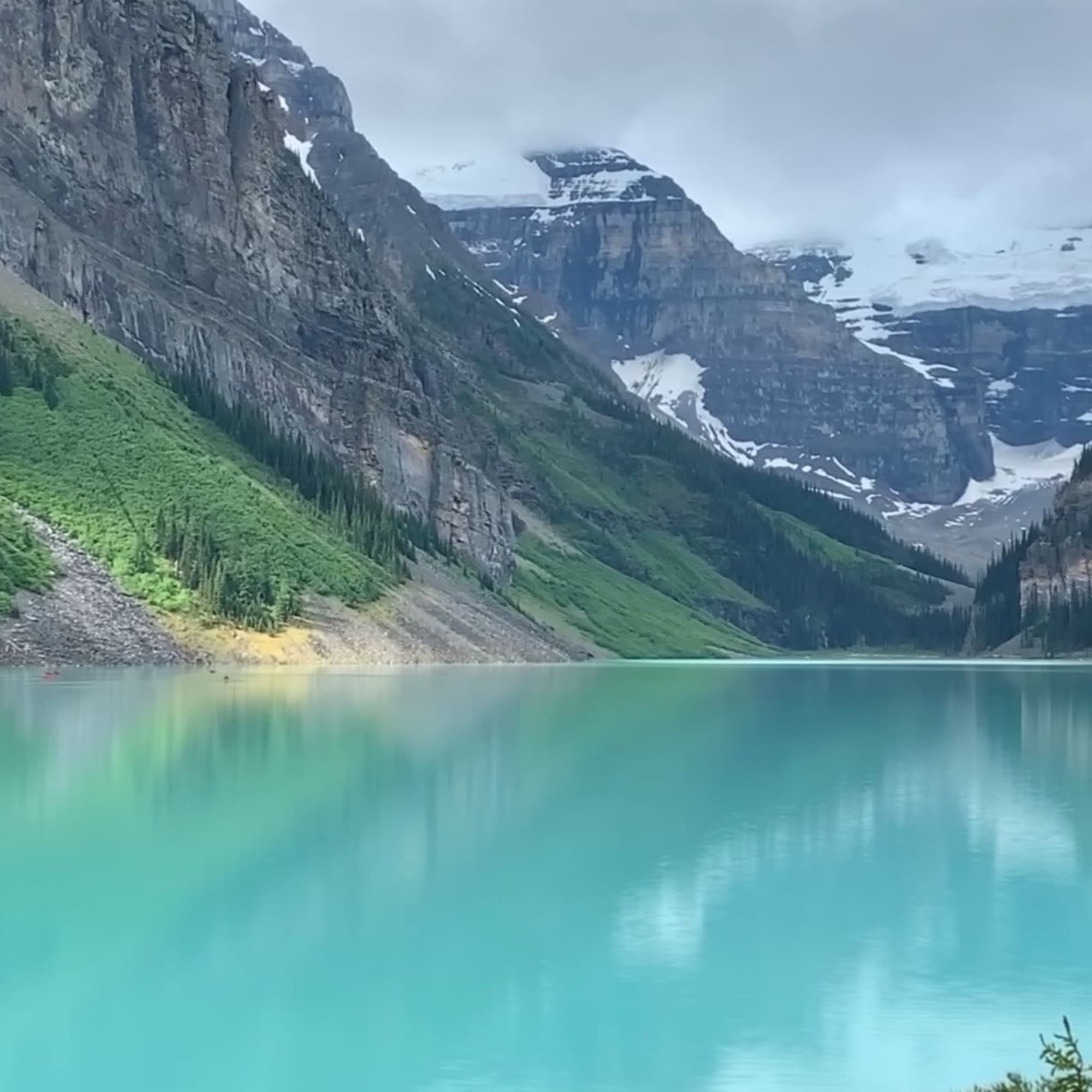 A vibrant turquoise lake is surrounded by lush greenery and snow-capped mountains under a cloudy sky.