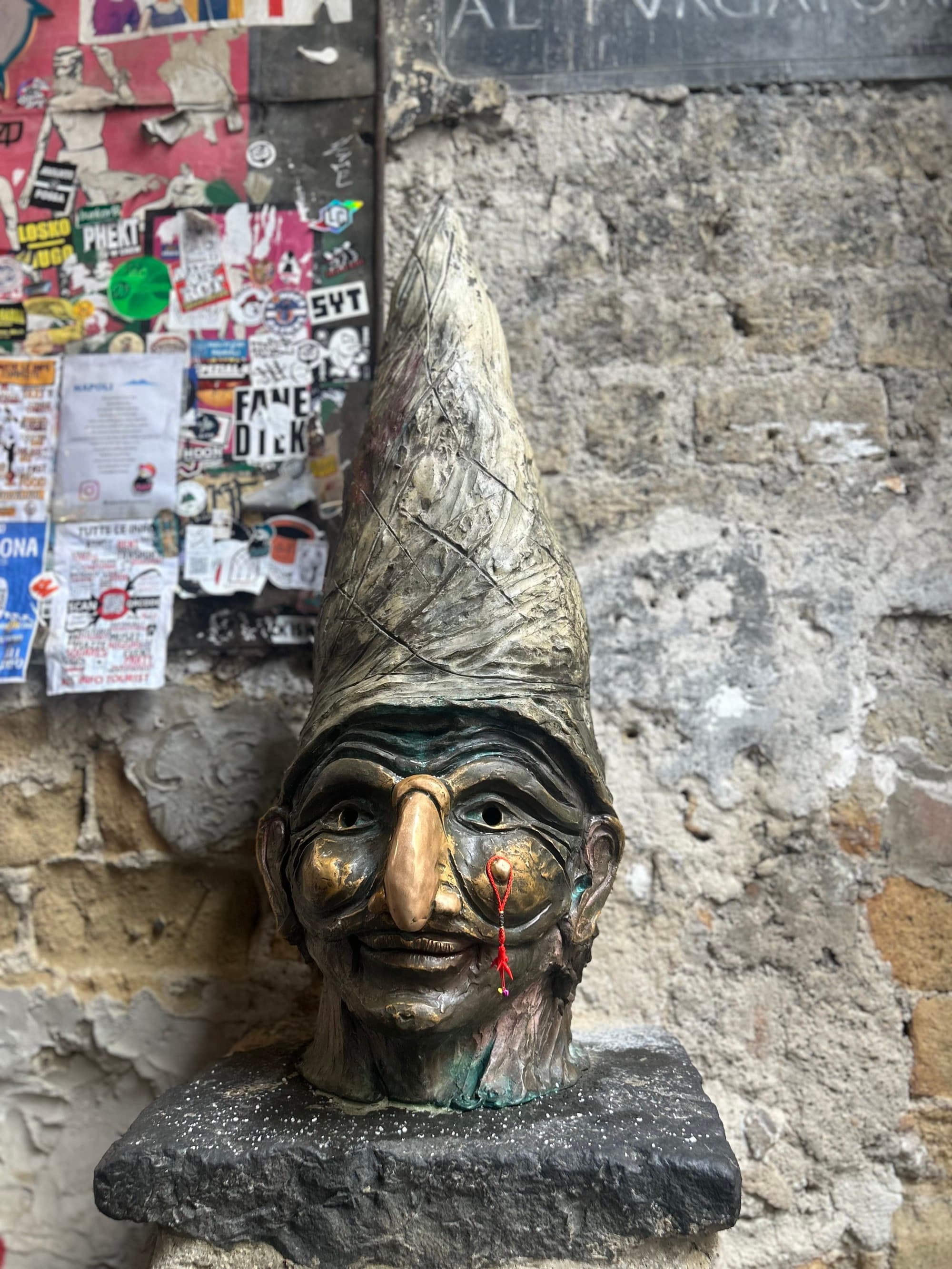 A statue with a pointed, tall hat with a red thread hanging from the cheek, against a stone wall with small notes attached.