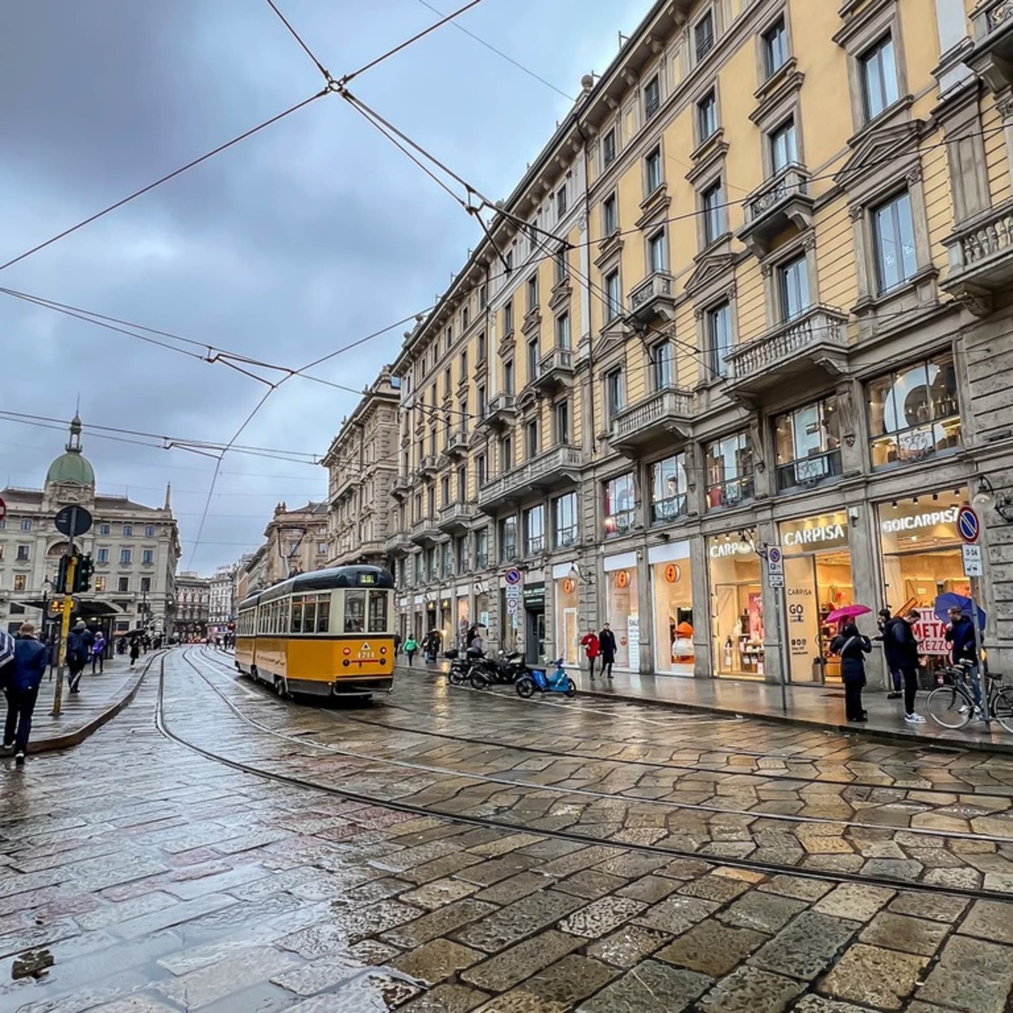 A wet urban street scene with a yellow tram, pedestrians, and classic architecture under an overcast sky