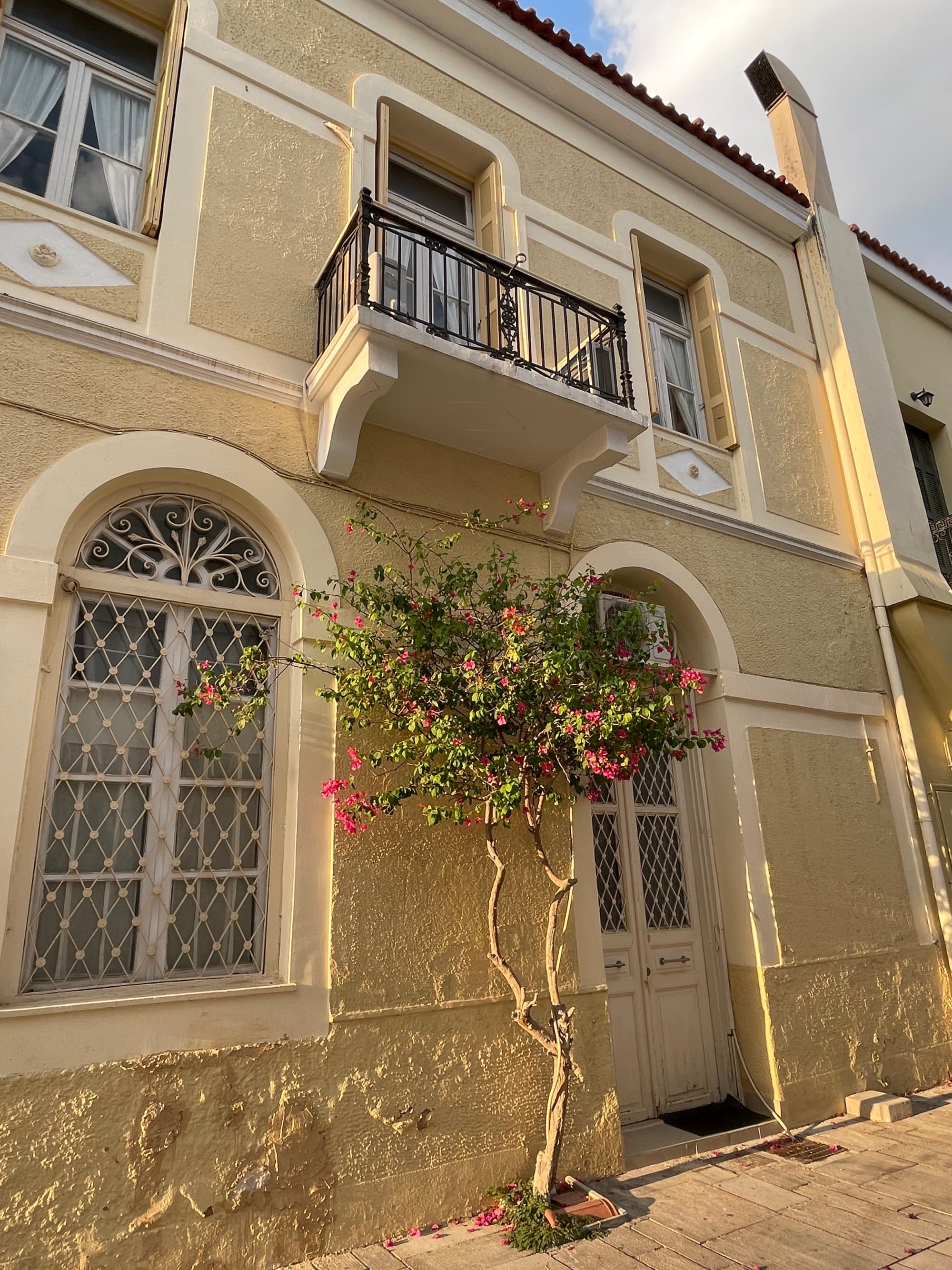 The image shows the exterior of a beige building with decorative windows, a small balcony and flowering plants.