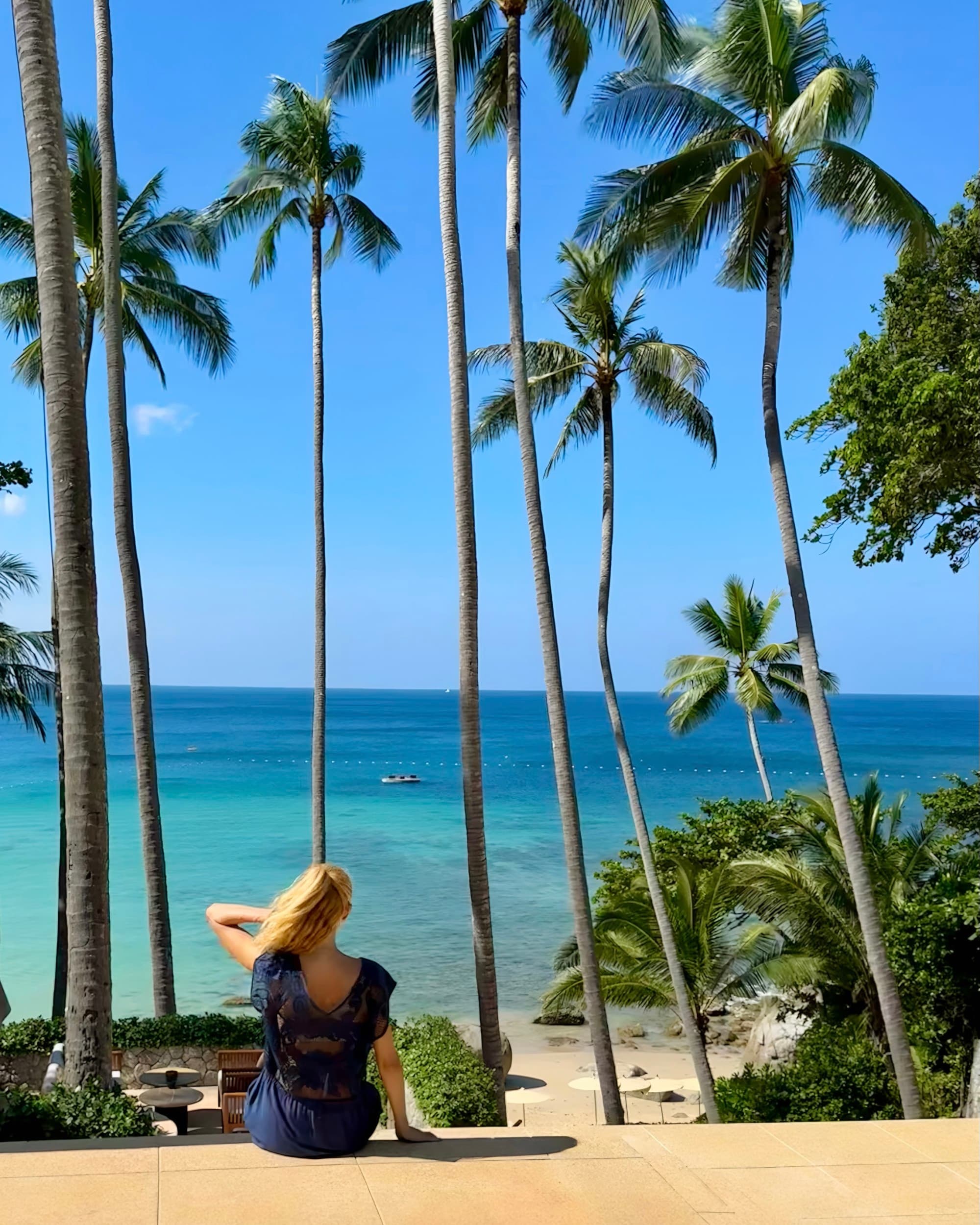 The image shows a person sitting on a bench looking out at a tropical beach with tall palm trees and clear blue water.