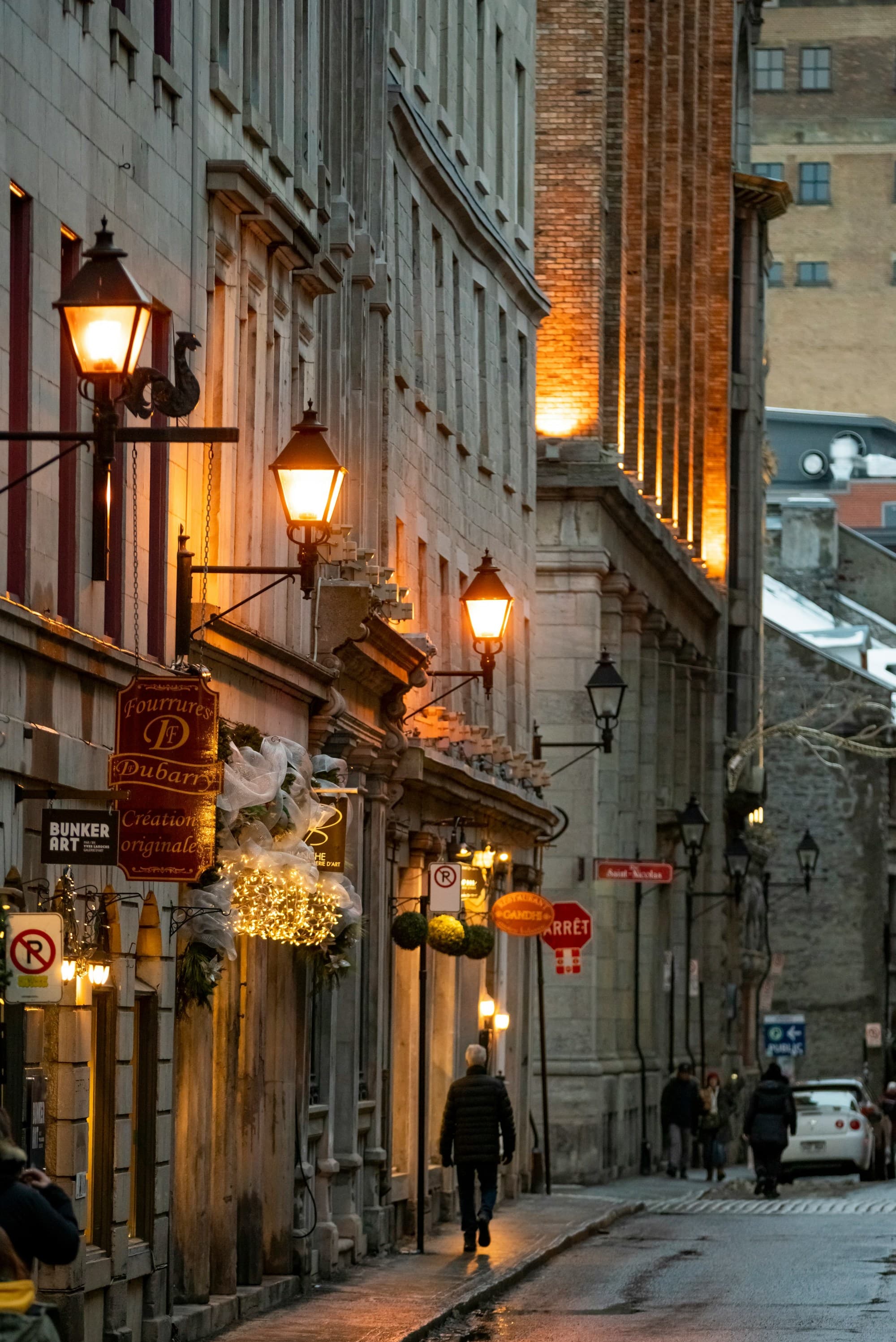 Street lamps warmly illuminate a narrow street lined with old buildings at dusk.