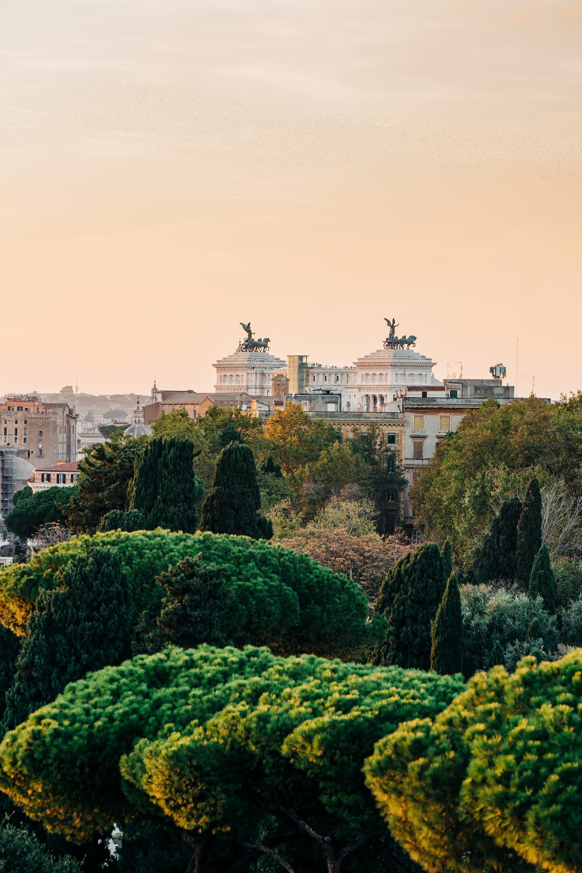 The image shows a beautiful cityscape at dusk, with buildings and statues silhouetted against a pastel sky and lush greenery in the foreground.