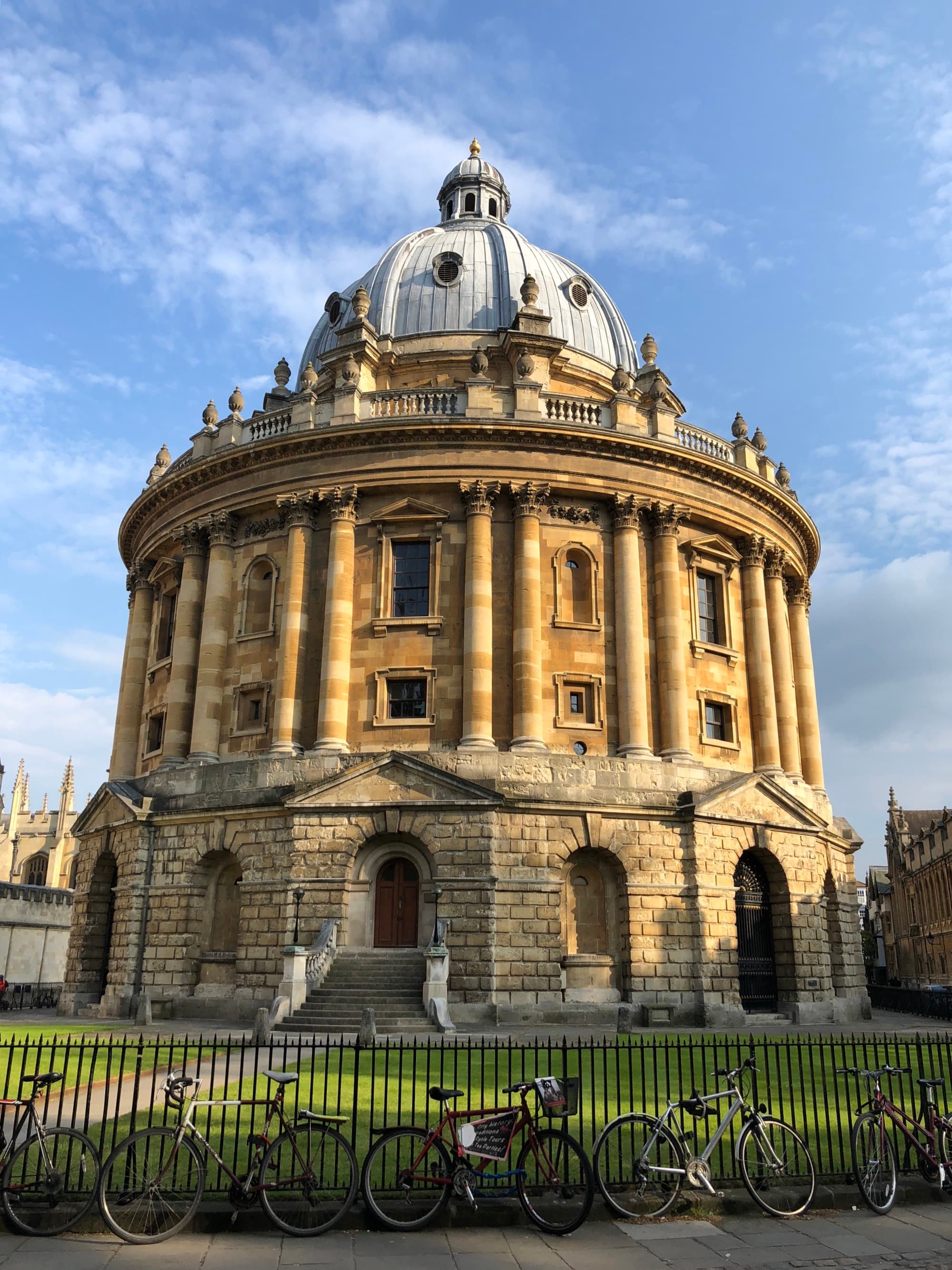 A view of a large dome building with ornate architexture and an iron gate surrounding it, known as the Radcliffe Camera, during the day.