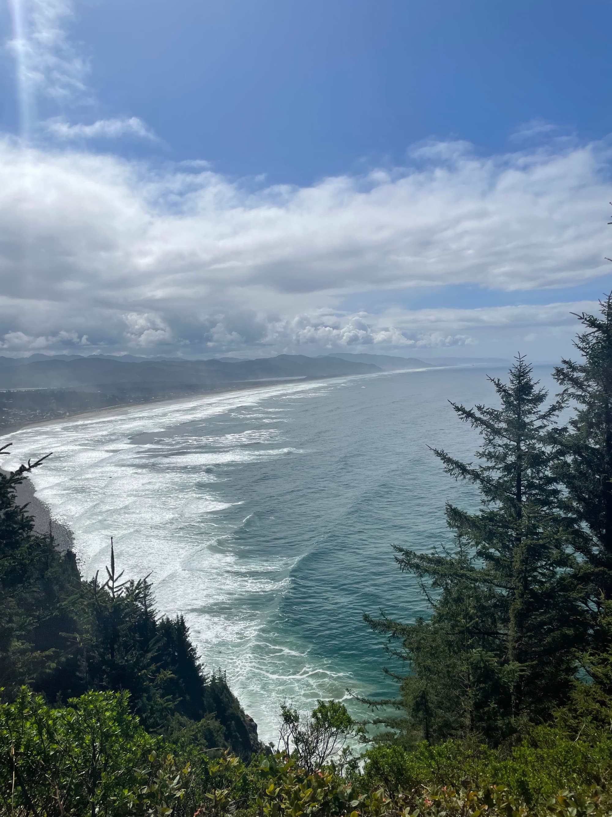 Sunlight filters through clouds, highlighting the ocean and forested cliffs in a scenic coastal landscape.