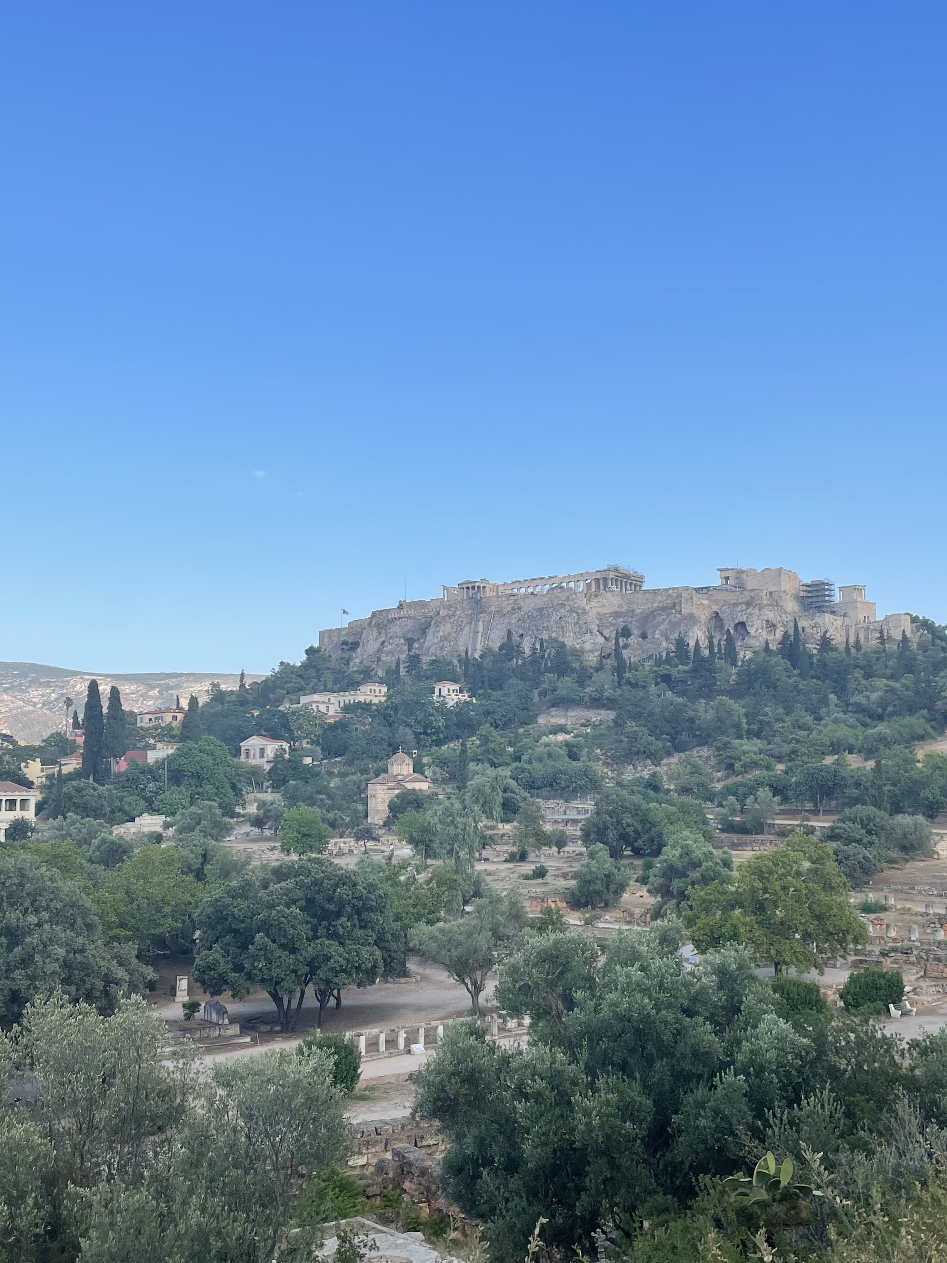 The Acropolis on a hilltop in the distance with green trees below it.