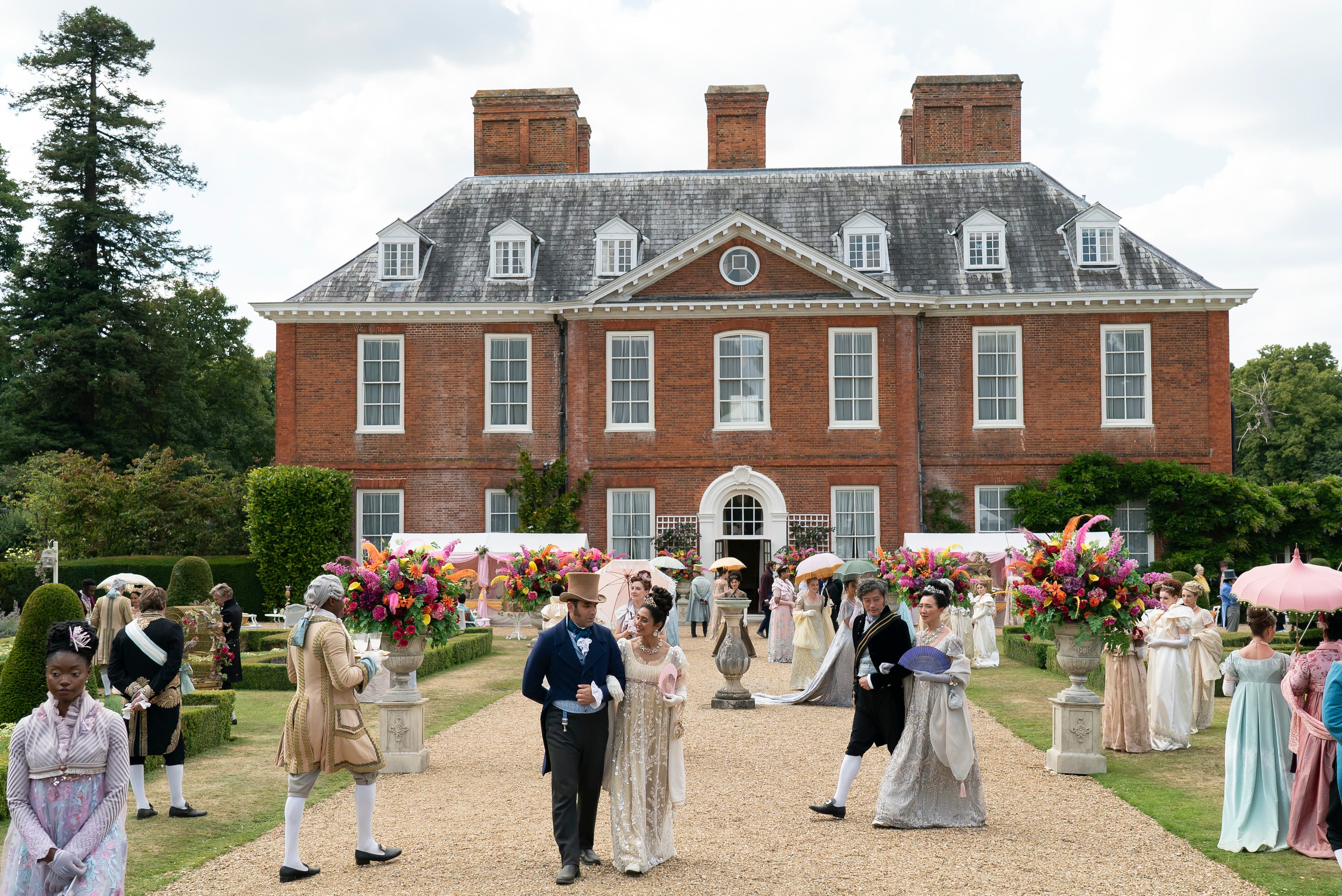 The image depicts a a view of a traditional stately home with a gravel path and beautiful gardens ini the UK with people walking in traditional, elegant outfits from the 19th century