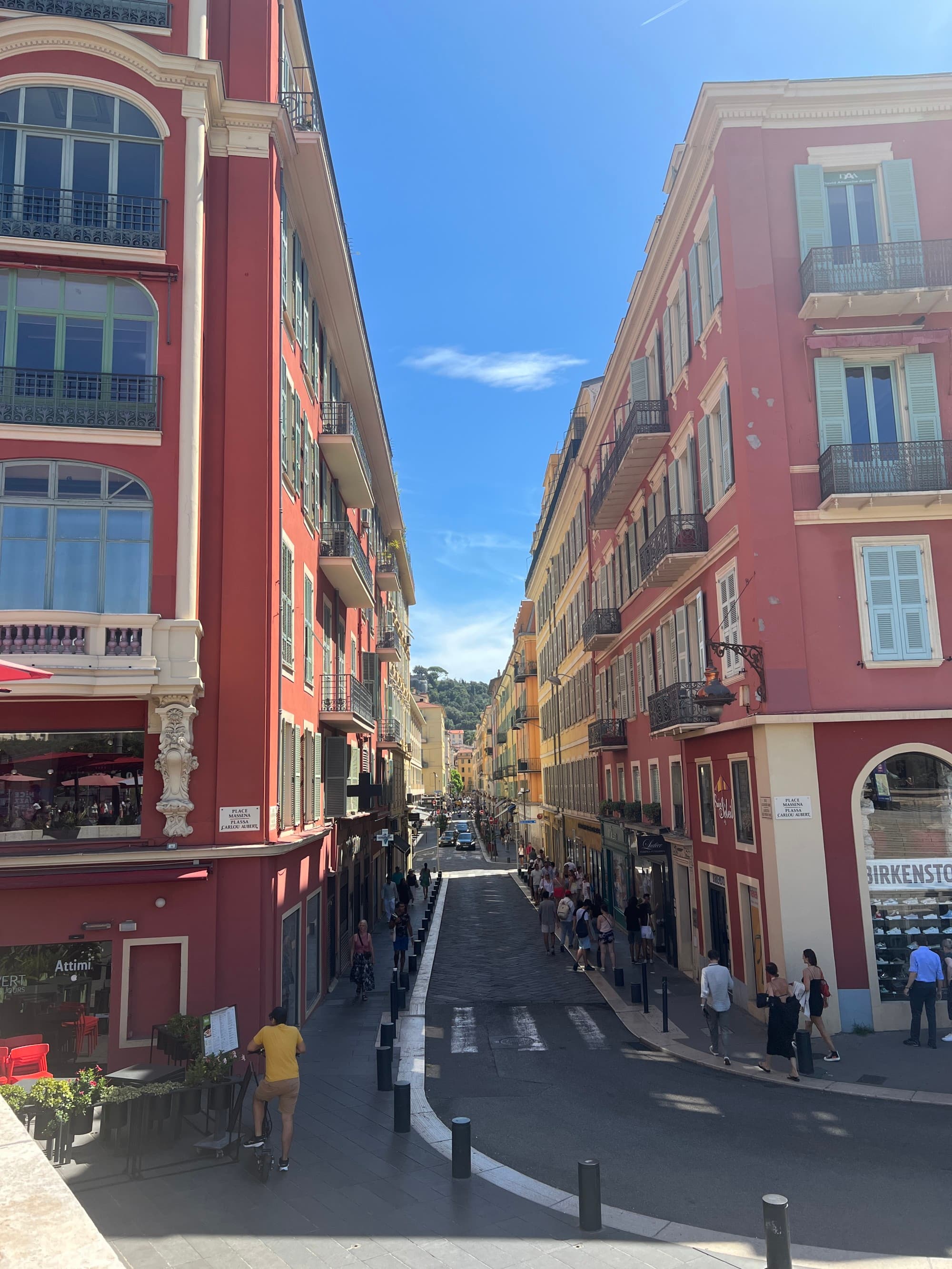 A view down a city street with pedestrians walking and maroon buildings with yellow trim on either side and a blue sky above.