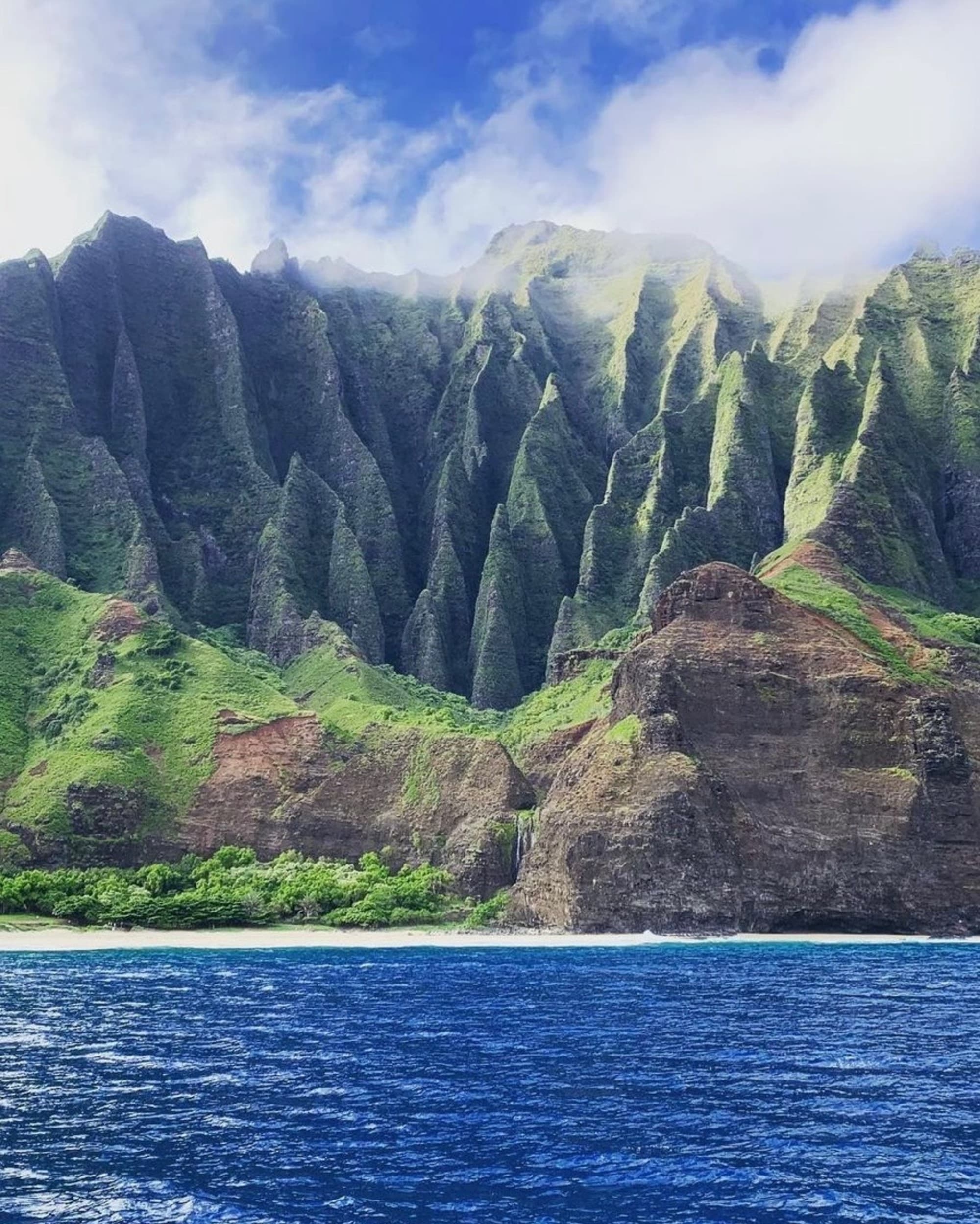 A series of tall, green, ridged cliffs overlooking a calm body of water under a cloudy sky.