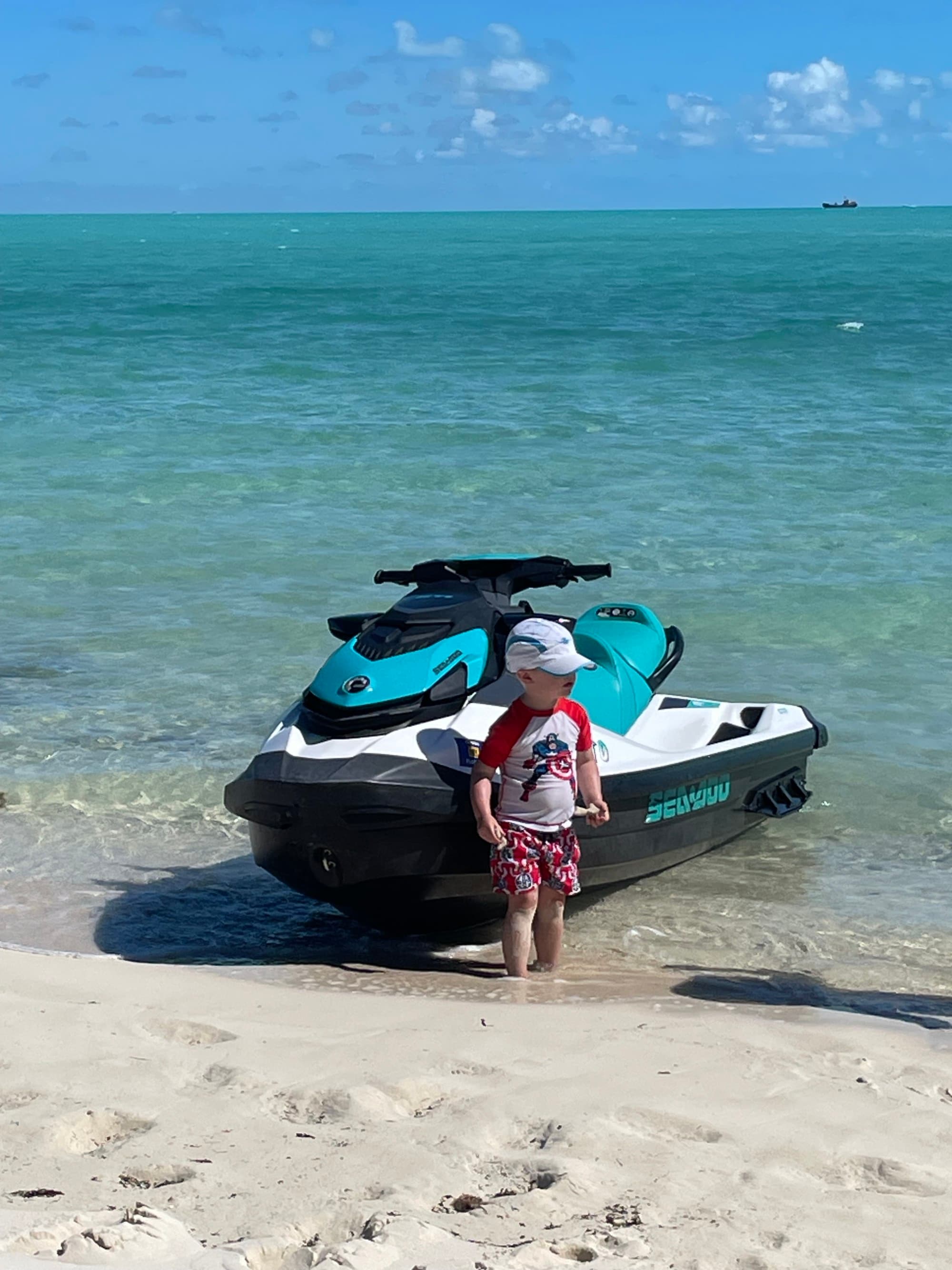 The image shows a person standing next to a blue and white jet ski on a sandy beach with the ocean in the background.