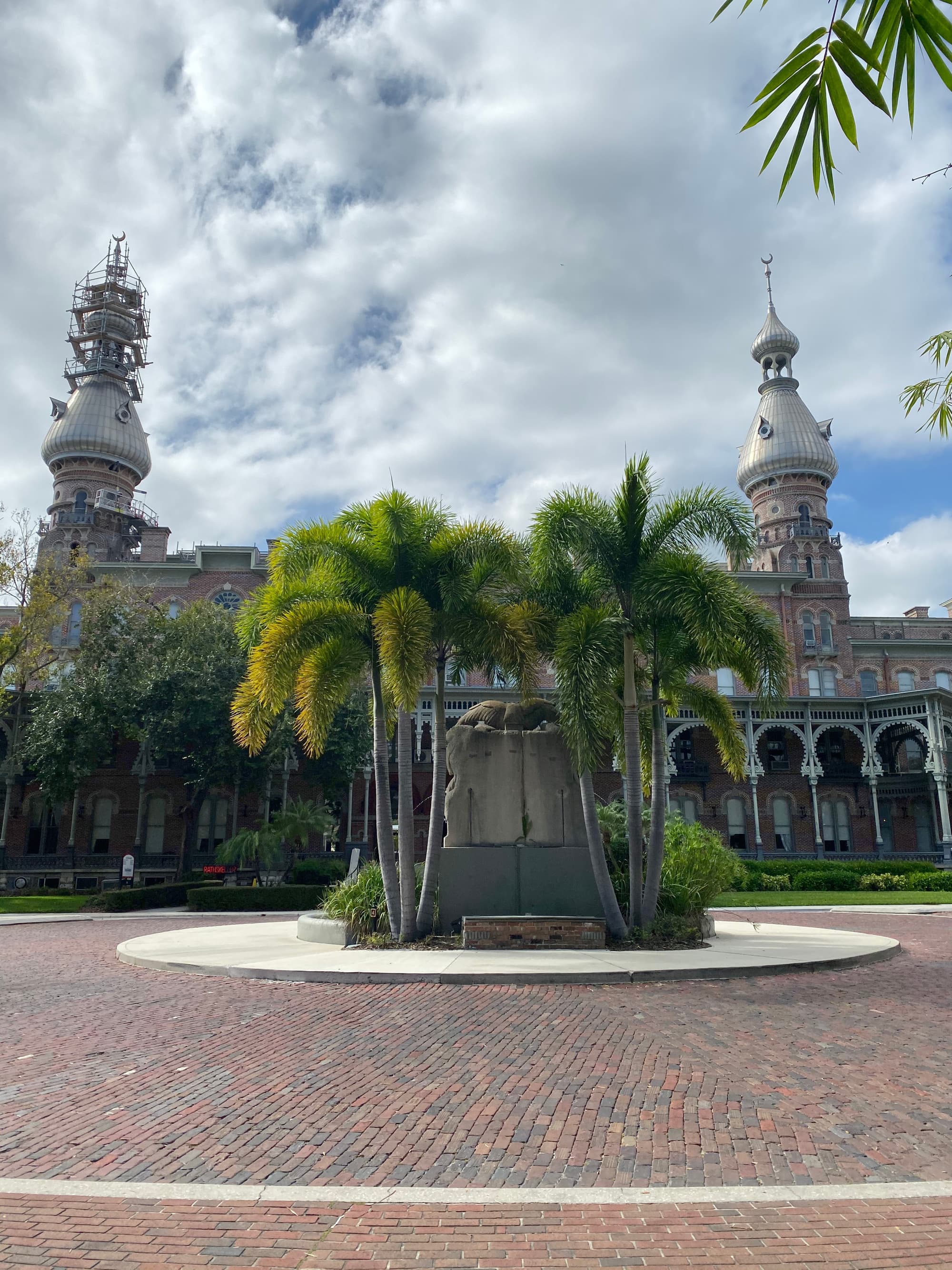 An outdoor scene with a brick pathway, palm trees, and a building with a spire in the background.