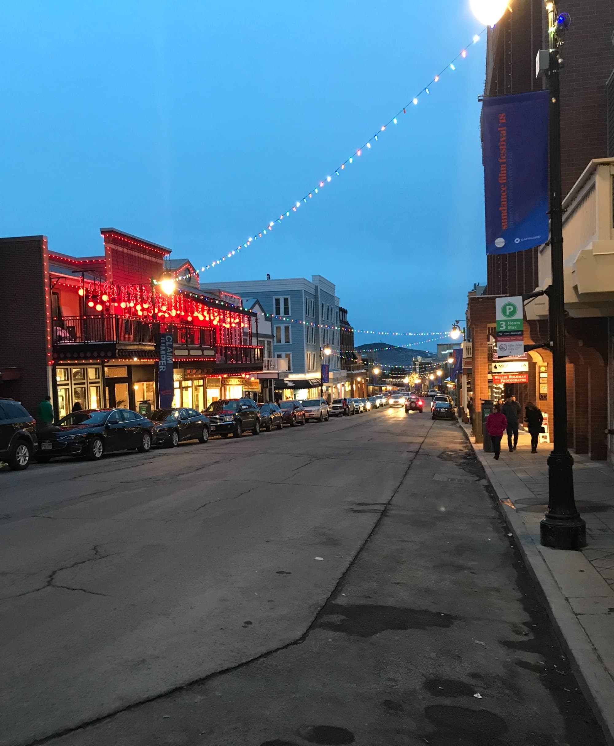 A view of Old Mainstreet at dusk, a paved road with lit up buildings on either side and cars parked out front.