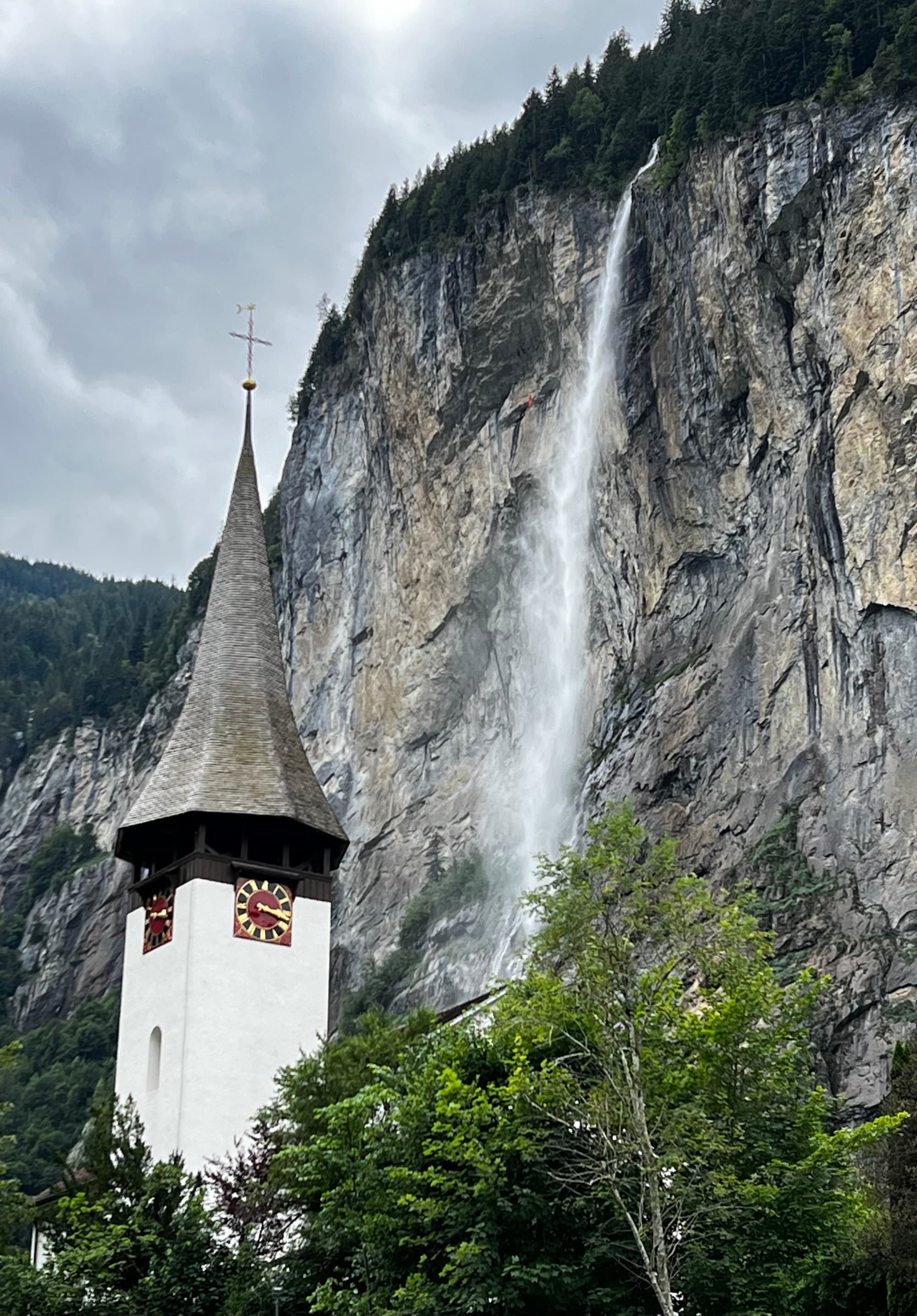 A church with a pointed steeple and clock stands against a backdrop of a tall cliff with a cascading waterfall.