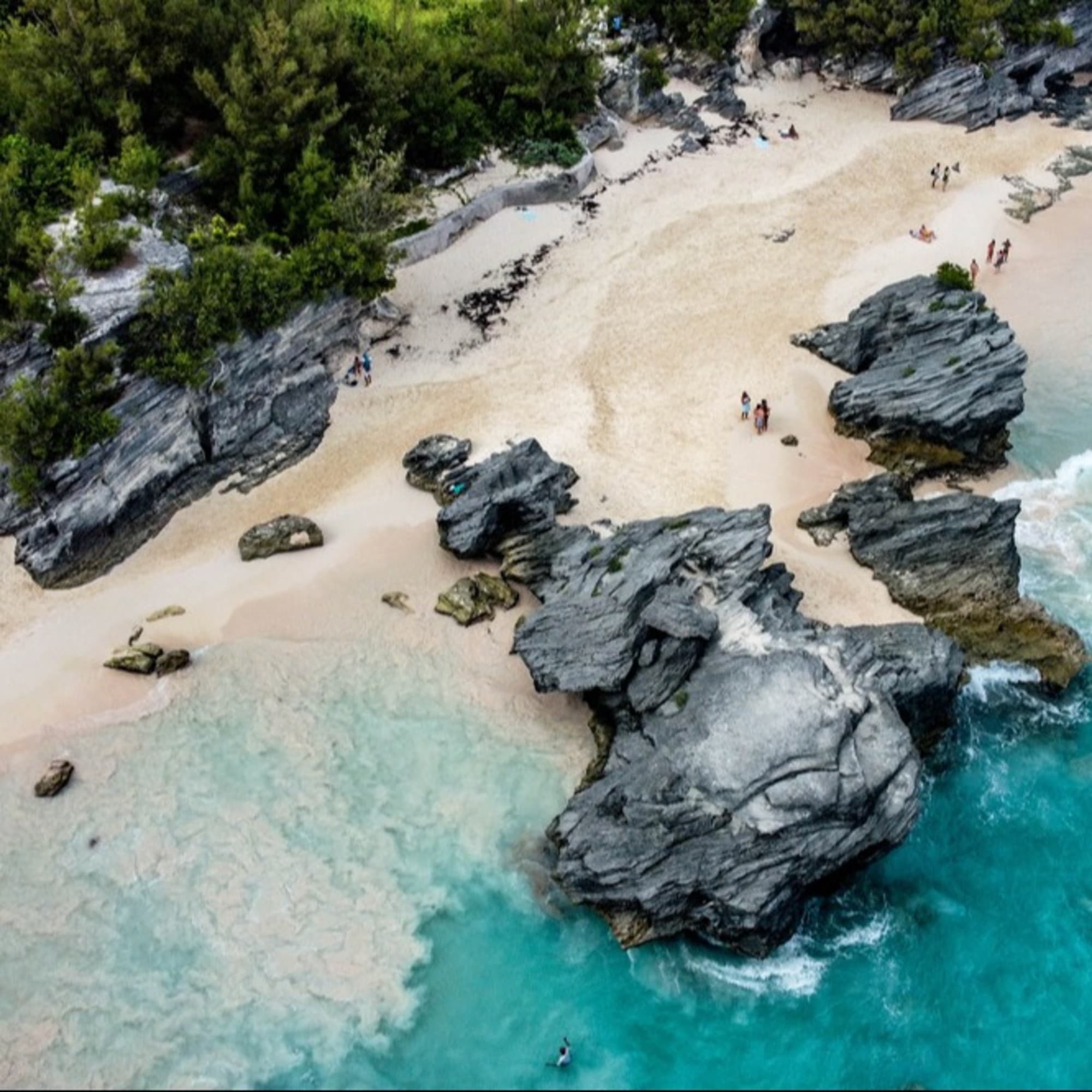 An aerial view of a beach with turquoise waters, large rocks, and people scattered on the sand.