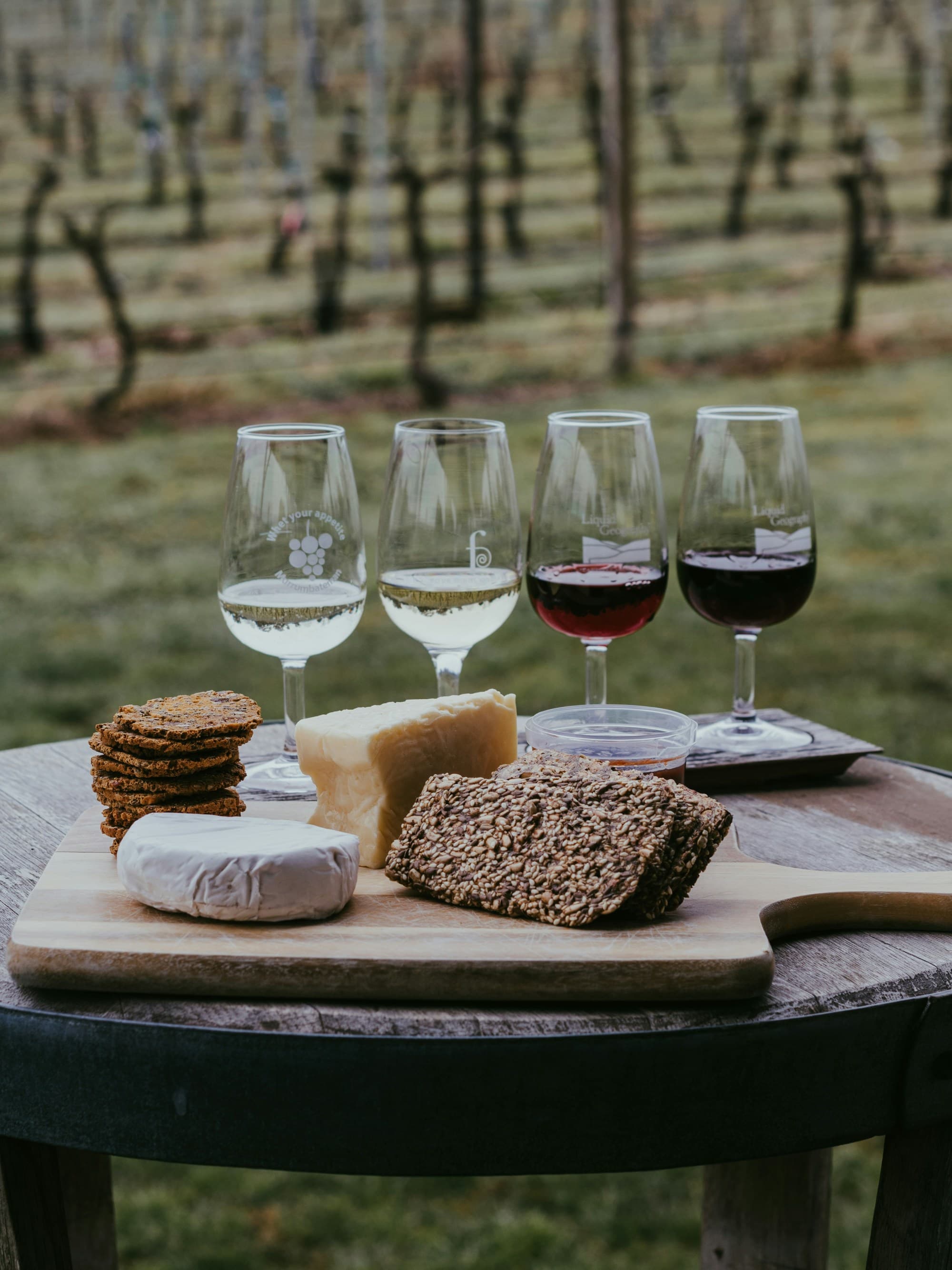 A selection of cheeses and cookies on a wooden board is paired with four glasses of wine, set against a vineyard backdrop.