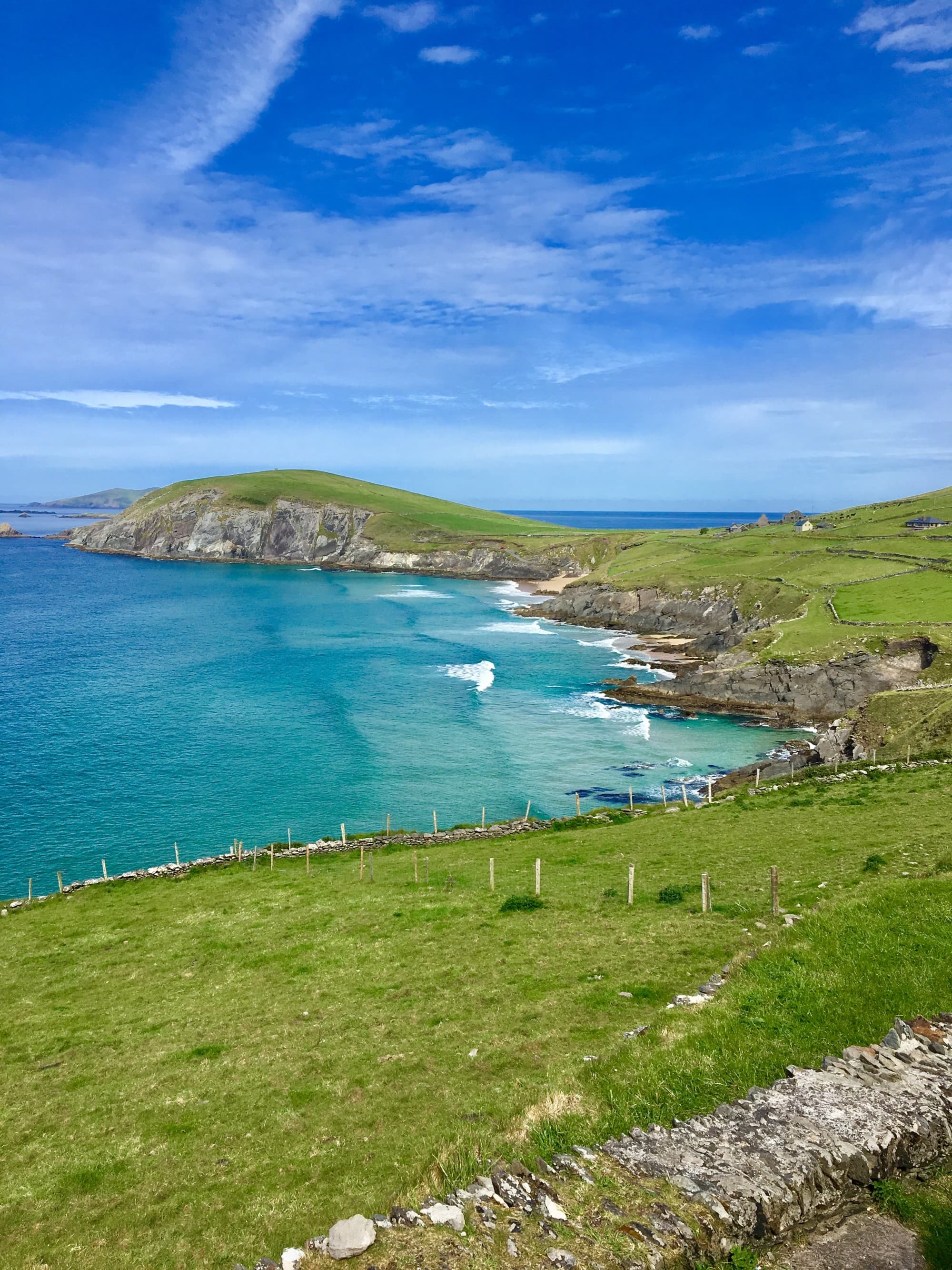 The image shows a scenic coastal landscape with lush green fields, a clear blue sky and a tranquil sea.